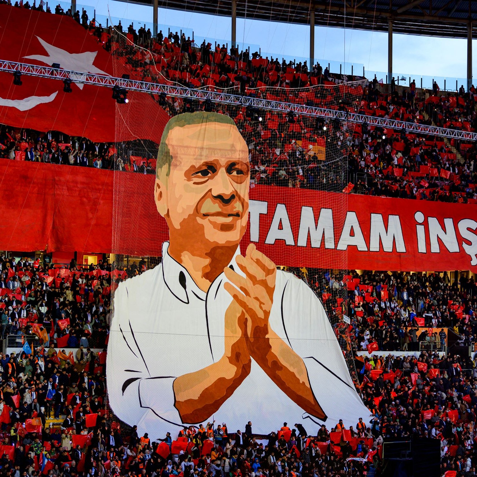 La imagen muestra un gran despliegue en un estadio, donde se aprecia una multitud de personas sosteniendo banderas rojas. En el centro, destaca un enorme cartel que presenta la figura de una persona aplaudiendo, acompañado de la frase "Tamam İnşallah", que se traduce como "Está bien, si Dios quiere." El ambiente parece ser festivo y de apoyo a esta figura, probablemente un líder político, en un evento significativo.