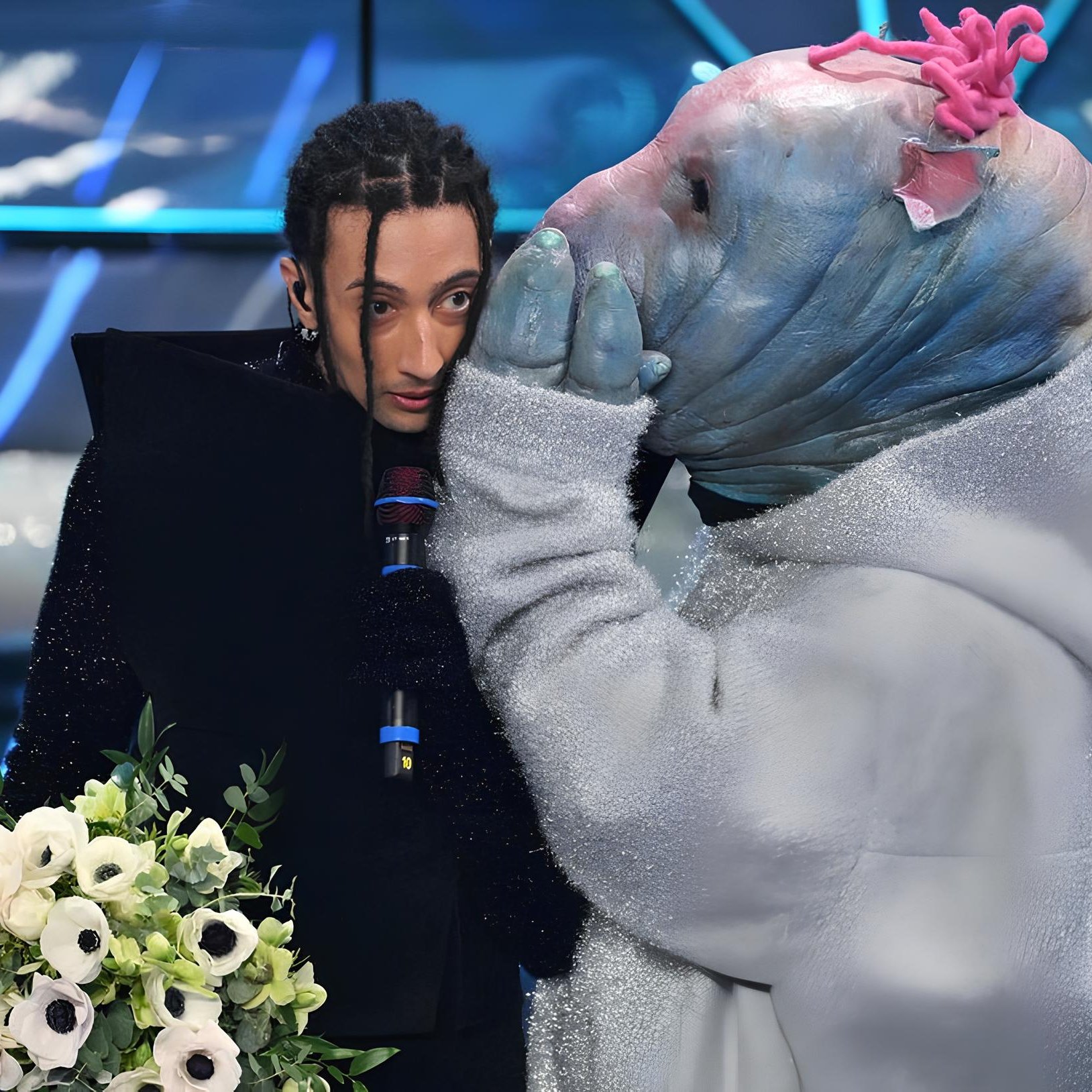 L'immagine mostra due figure in un ambiente scenico. Una persona, vestita con abiti scuri e con capelli lunghi e intrecciati, tiene un microfono e un mazzetto di fiori. Accanto a lei c'è un personaggio mascherato, che indossa un costume piumato e una maschera con tratti insoliti. Il personaggio sembra sussurrare all'orecchio della persona, creando un'atmosfera di intimità o mistero. Sullo sfondo si possono vedere luci blu e decorazioni moderne, tipiche di uno spettacolo televisivo.