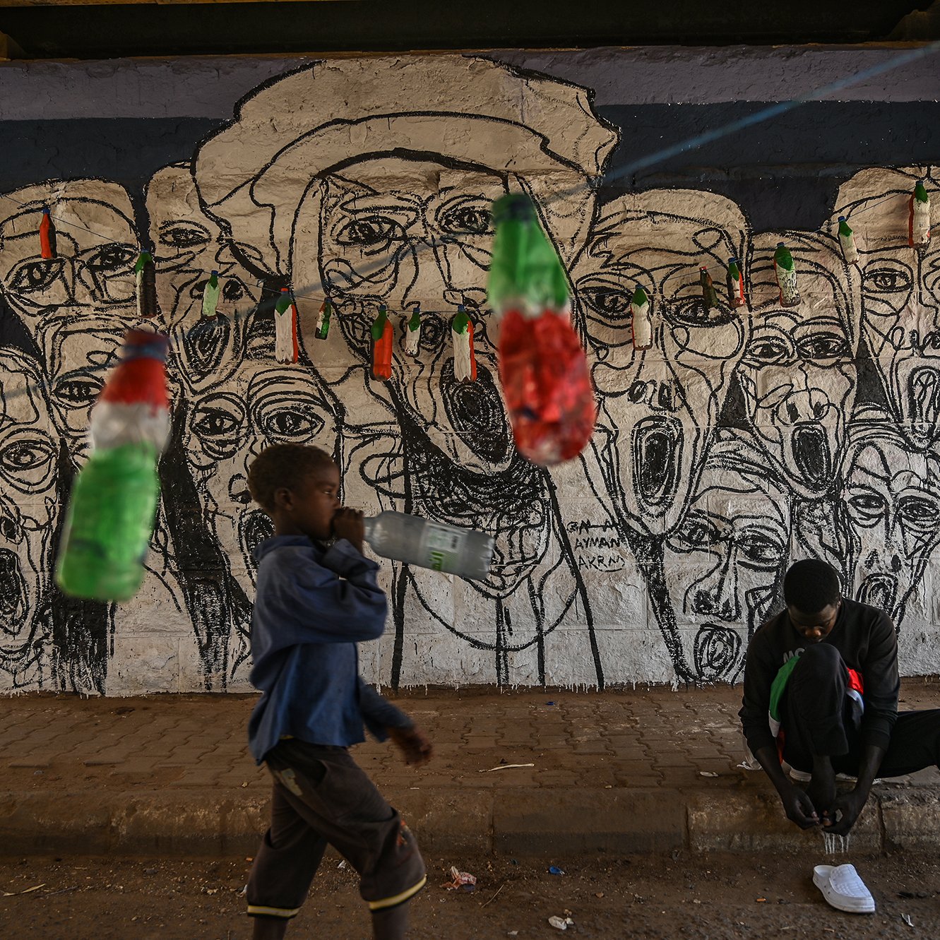 L'image montre un mur peint en arrière-plan, où se trouvent des visages aux traits exagérés, tous avec des expressions de cri, créant une atmosphère vibrante et expressive. Devant ce mur, un enfant marche en tenant une bouteille, tandis qu'un autre individu est assis au sol, occupé à lacer ses chaussures. Des bouteilles colorées, probablement en plastique, sont suspendues au-dessus d'eux, ajoutant une touche de couleur à la scène. Cette image reflète un environnement urbain dynamique, mêlant art de rue et vie quotidienne.