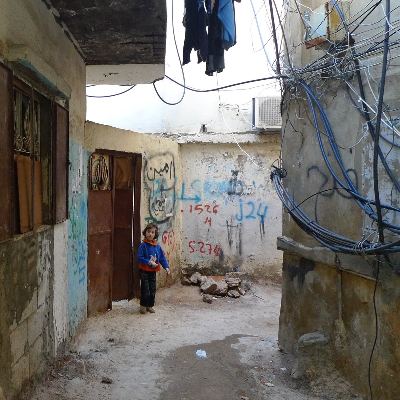 الصورة تظهر زقاقًا ضيقًا بين مباني قديمة، حيث تشاهد جدرانًا مغطاة برسوم جدارية وكتابات متنوعة. في أحد الجدران، هناك ملصقات وأوراق مثبتة. كما تظهر الأسلاك الكهربائية متدلية من فوق. في نهاية الزقاق، يقف طفل يرتدي قميصًا أزرق، يبدو فضولياً وهو ينظر إلى الكاميرا. الأجواء عامة تعكس حالة من الفقر والبساطة.