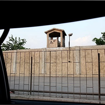 L'image montre un mur de béton surmonté d'une petite structure, probablement un poste de garde, avec un toit en pente. Au premier plan, on peut voir une grille métallique qui sépare l'intérieur de la scène extérieure. En arrière-plan, des arbres verdoyants apparaissent, ajoutant une touche de nature à l'environnement urbain. La lumière de la fin de journée crée une atmosphère calme.