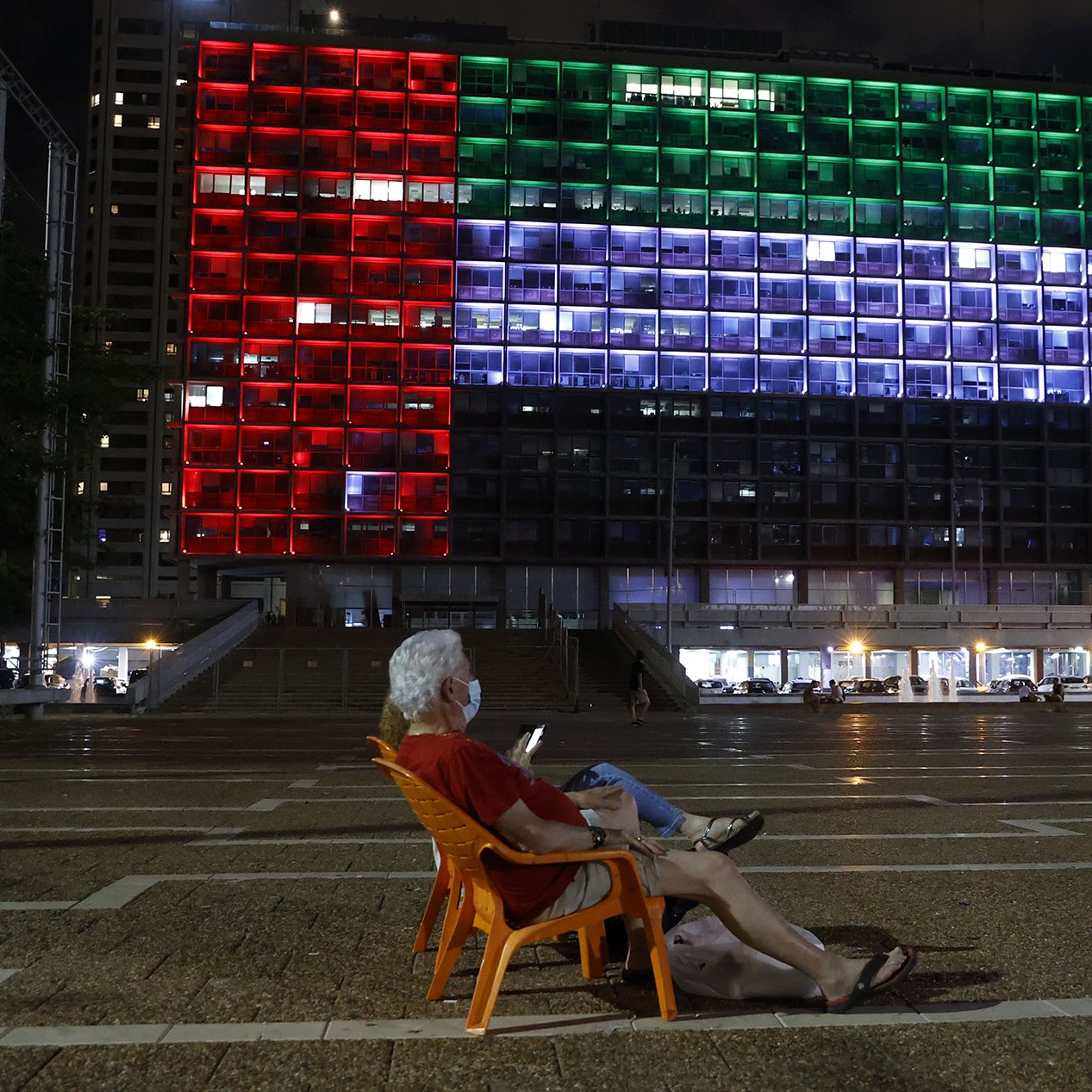 L'image montre une scène nocturne dans une ville. Au premier plan, un homme âgé est assis sur une chaise orange, en train de regarder son téléphone. En arrière-plan, un bâtiment imposant est illuminé avec des lumières de différentes couleurs, formant un motif qui ressemble à un drapeau. Les couleurs rouge, vert et bleu s'alternent sur la façade du bâtiment, créant une ambiance vibrante. D'autres personnes se déplacent dans l'espace public, ajoutant de la vie à la scène.