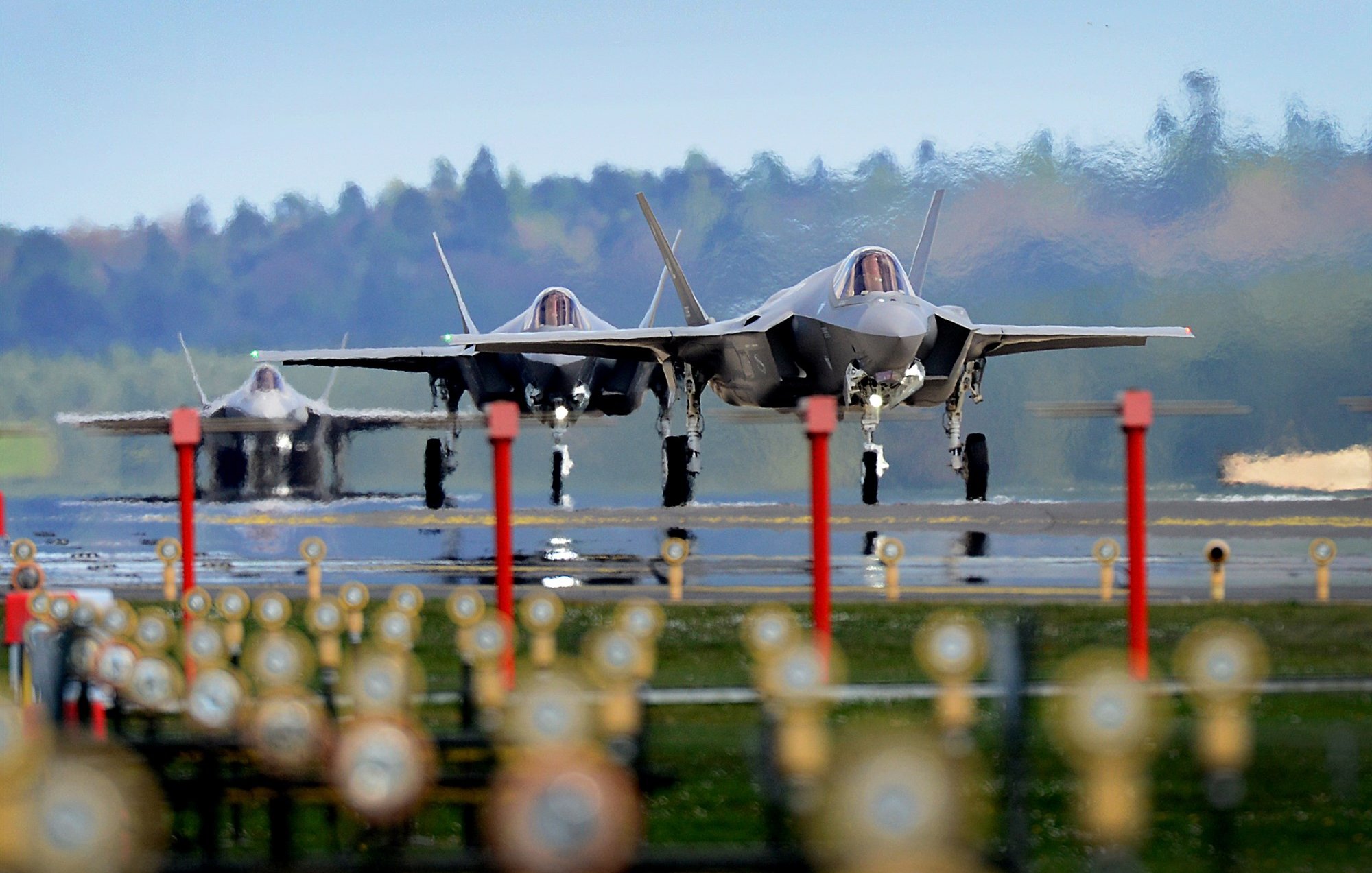 The image features three F-35 fighter jets on an airstrip, showcasing their sleek and modern designs. The jets are positioned closely together, and the background features a blurred green landscape, indicating a rural setting. The foreground includes several lights lining the runway, which adds a sense of depth to the scene. The overall atmosphere appears dynamic and focused on military aviation.