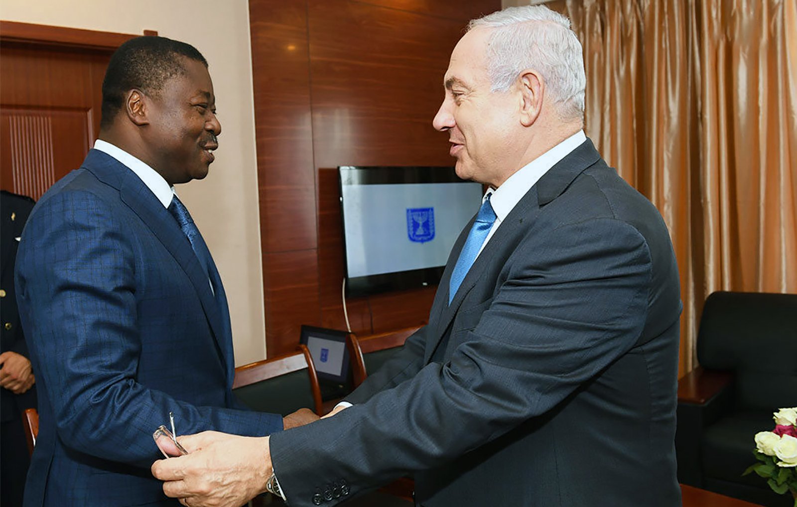 The image depicts two men engaging in a handshake in a formal setting. One man is dressed in a blue suit and tie, while the other is in a dark suit and tie. They appear to be smiling and are facing each other, suggesting a cordial meeting or discussion. The background includes a decorative setting with drapes and some furniture, indicating a professional atmosphere.