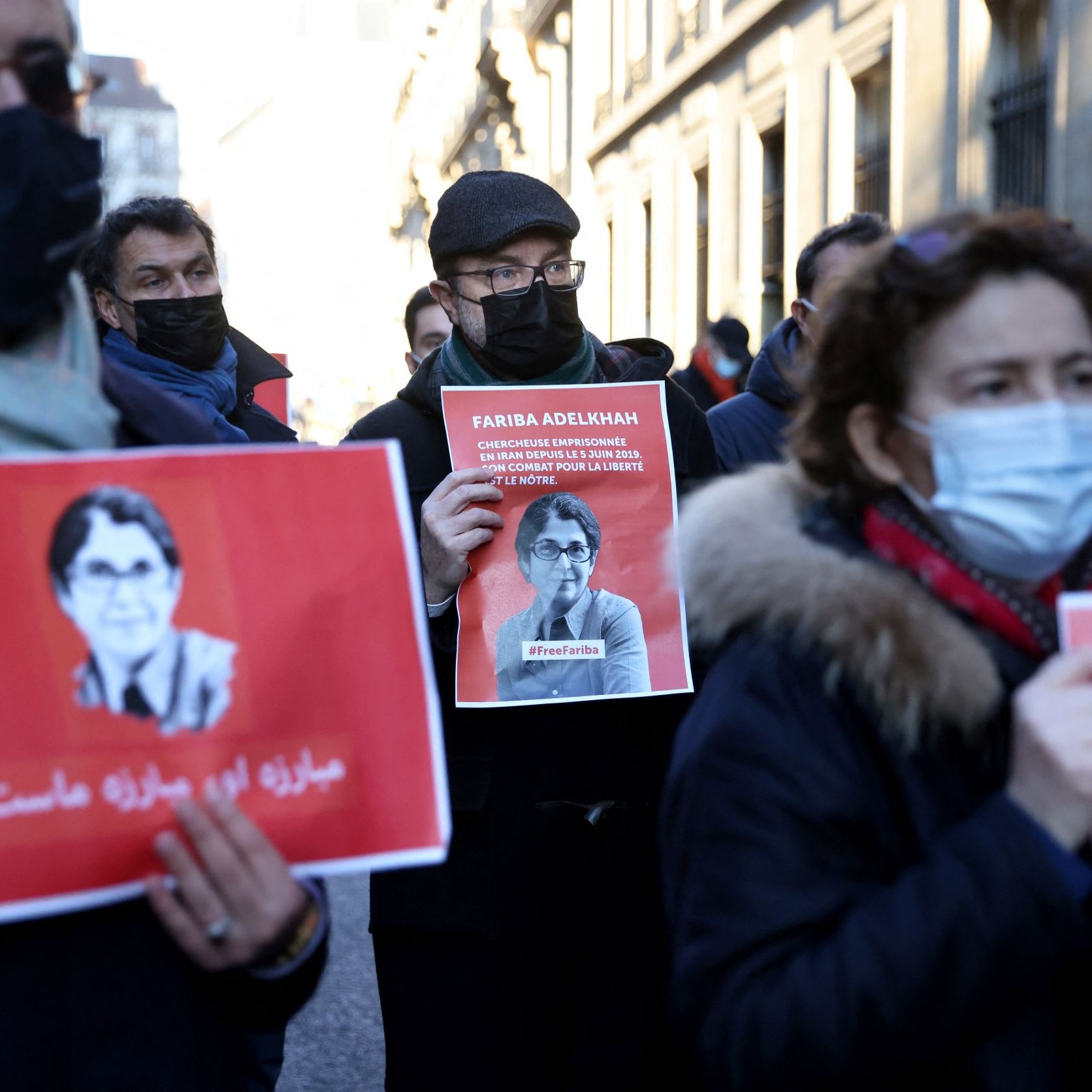 L'image montre un groupe de personnes participant à une manifestation. Elles tiennent des pancartes avec l'image et le nom d'une personne, Farida Adlénah, accompagnées de slogans. Les manifestants portent des masques, probablement en raison de préoccupations sanitaires. L'ambiance semble être solennelle et engagée, avec des visages sérieux. Les arrière-plans montrent des bâtiments qui pourraient avoir une architecture urbaine.