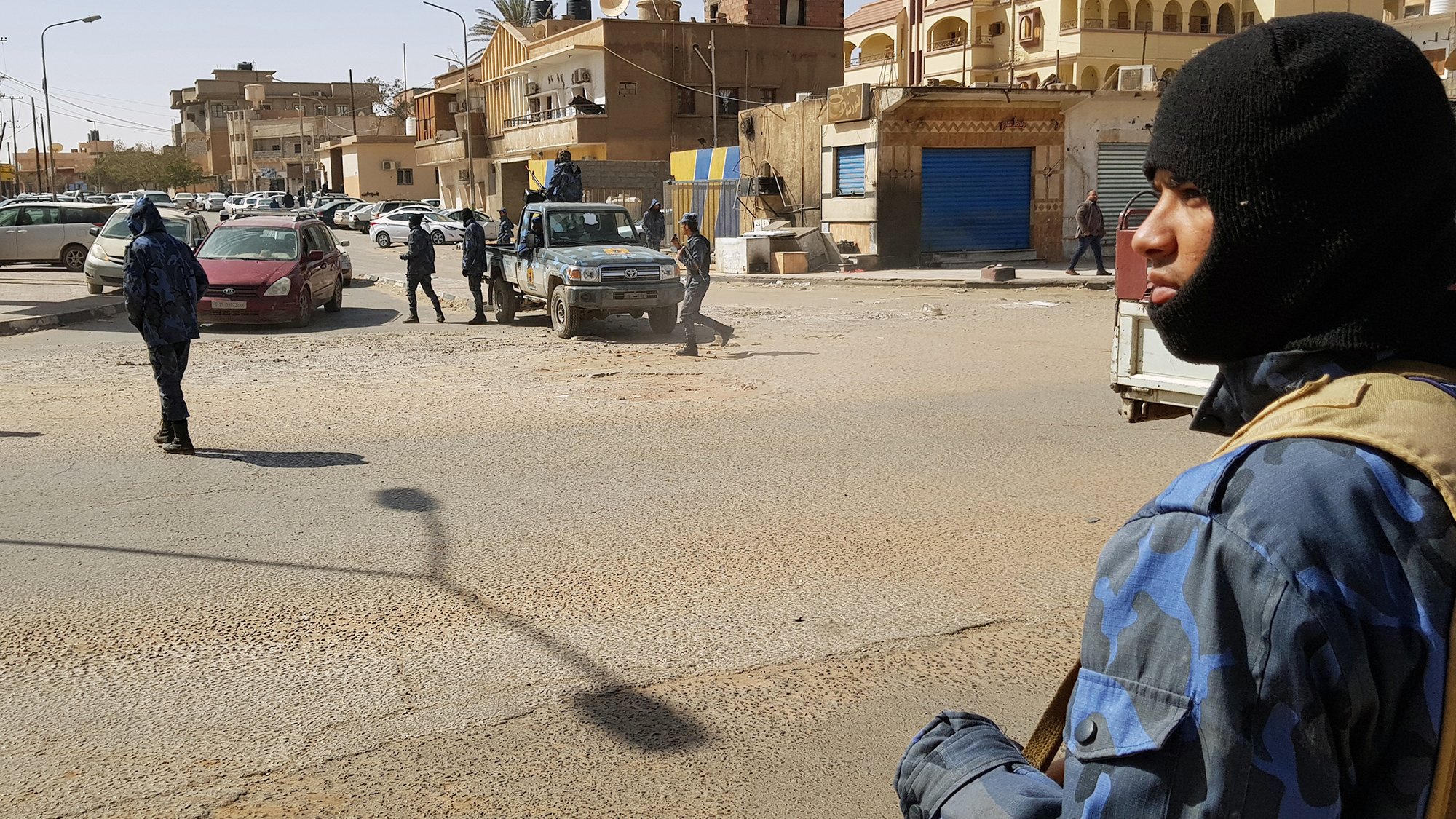 The image depicts a street scene in an urban environment where armed security personnel are present. One individual is prominently featured in the foreground, wearing a dark mask and tactical gear, holding a firearm. In the background, other security personnel are visible, some moving and others standing near vehicles, including a jeep. The street appears to be busy, with buildings lining either side and several parked cars. The setting suggests heightened security or law enforcement activity.