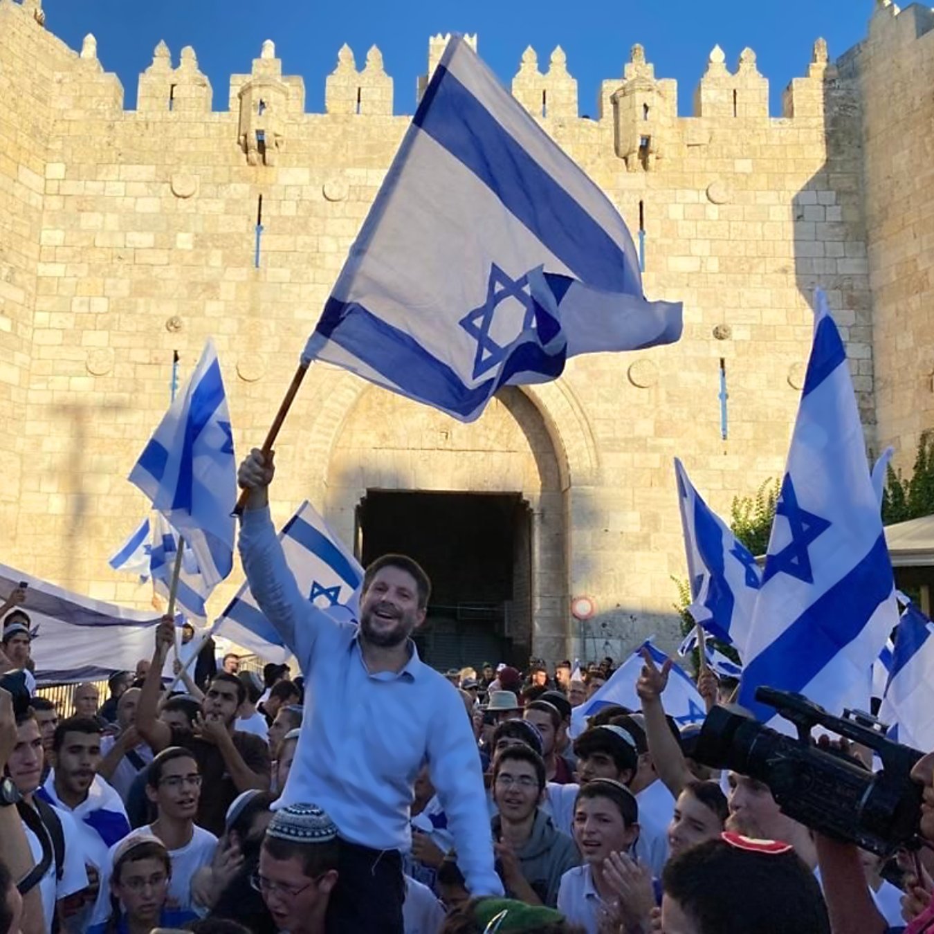تصویر نشان‌دهنده جمعیتی است که با پرچم‌های اسرائیل در حال جشن و شادی هستند. در میان این جمعیت، فردی در حال بلند کردن پرچم و ابراز شادی است. پس‌زمینه تصویر یک دروازه تاریخی و دیوار سنگی نمایان است که نشان‌دهنده یک مکان مهم است. افراد حاضر در تصویر لباس‌های مختلفی به تن دارند و فضای کلی عکس نشان‌دهنده شور و نشاط است.