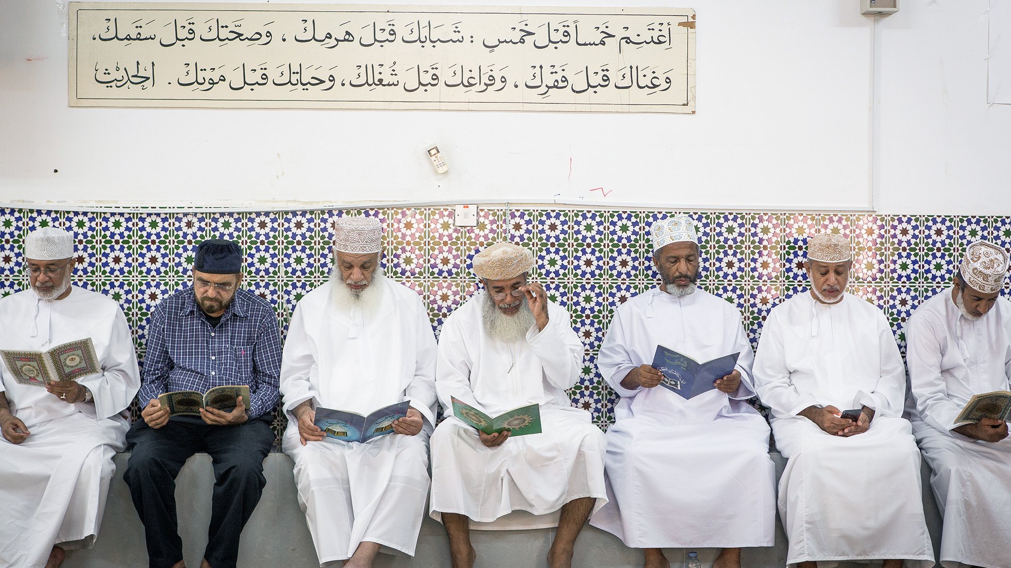L'image montre un groupe d'hommes assis en ligne, lisant des livres, probablement des copies du Coran. Ils portent des vêtements traditionnels, tels que des dishdashas et des couvre-chefs. En arrière-plan, on peut voir un mur décoré de carrelage coloré, sur lequel est inscrite une citation ou un texte. L'atmosphère semble calme et sereine, suggérant un moment de prière ou de méditation.