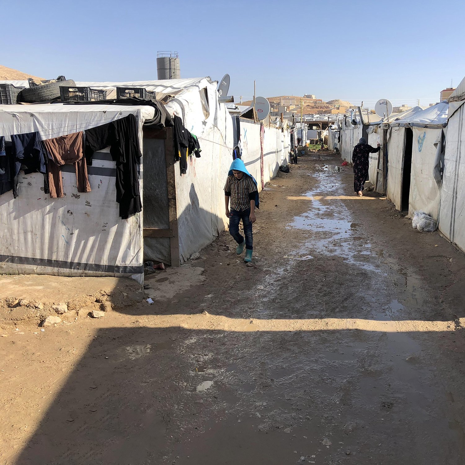 این تصویر نمایی از یک کمپ پناهندگان را نشان می‌دهد. در اینجا، چادرهای سفید و مشکی به صورت ردیفی در دو طرف یک مسیر خاکی قرار دارند. زمین به نظر می‌رسد کمی مرطوب و گل‌آلود است. چندین قطعه لباس بر روی چادرها آویزان شده‌اند و شخصی در حال حرکت در مسیر دیده می‌شود. در پس‌زمینه، ساختمان‌هایی در بالای تپه‌ها قابل مشاهده هستند. این منظره فضایی ساده و دلگیر را تداعی می‌کند.