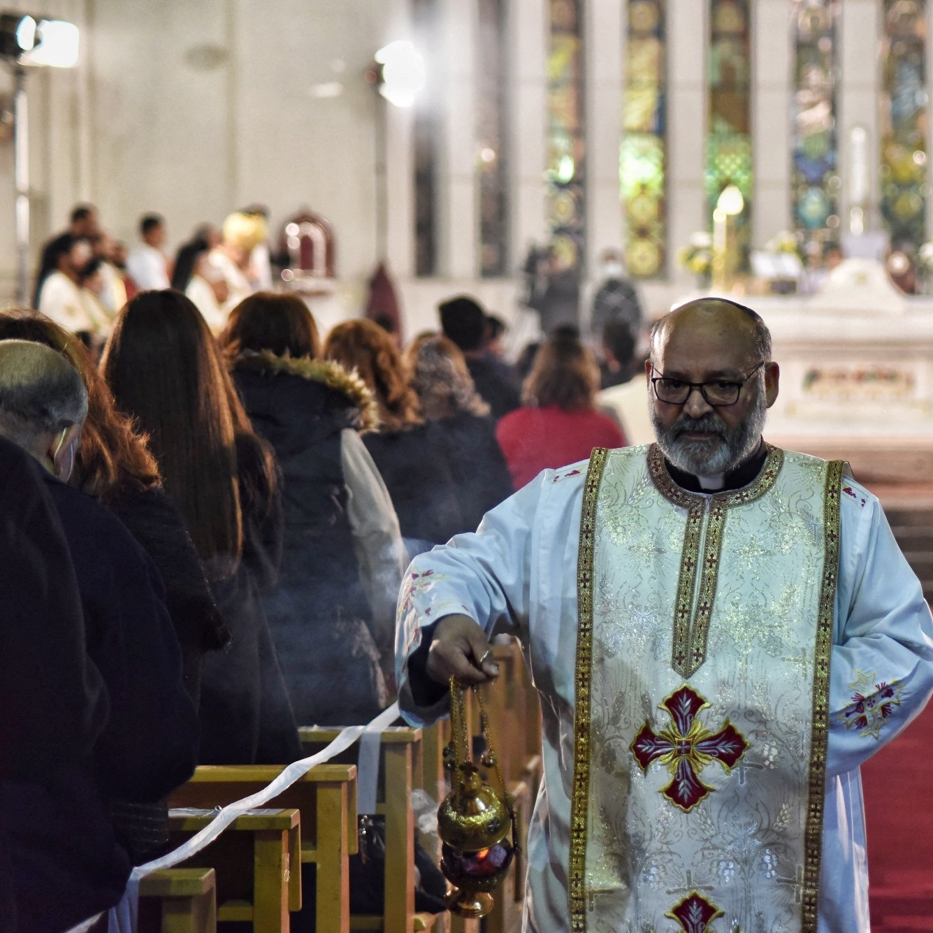 A priest in ornate vestments walks down an aisle in a church, surrounded by worshippers.