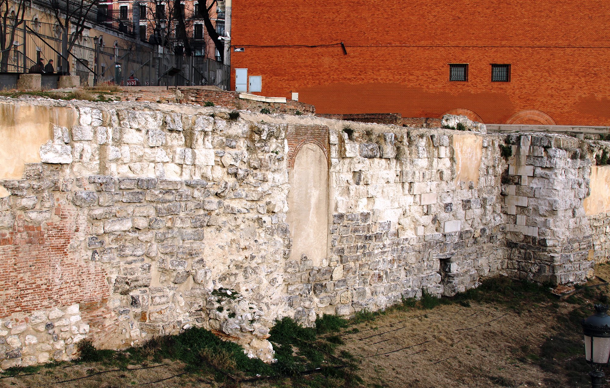 La imagen muestra una pared de piedra antigua, probablemente parte de una estructura histórica. La pared está compuesta de grandes bloques de piedra, algunos de los cuales están desgastados o dañados. En el centro, hay un espacio vacío en la pared que sugiere la existencia previa de una puerta o ventana. Al fondo, se puede ver un edificio de ladrillo rojo y un área de césped seco. Hay algunos restos de vegetación en la base de la pared, lo que añade un toque de naturaleza al entorno urbano.