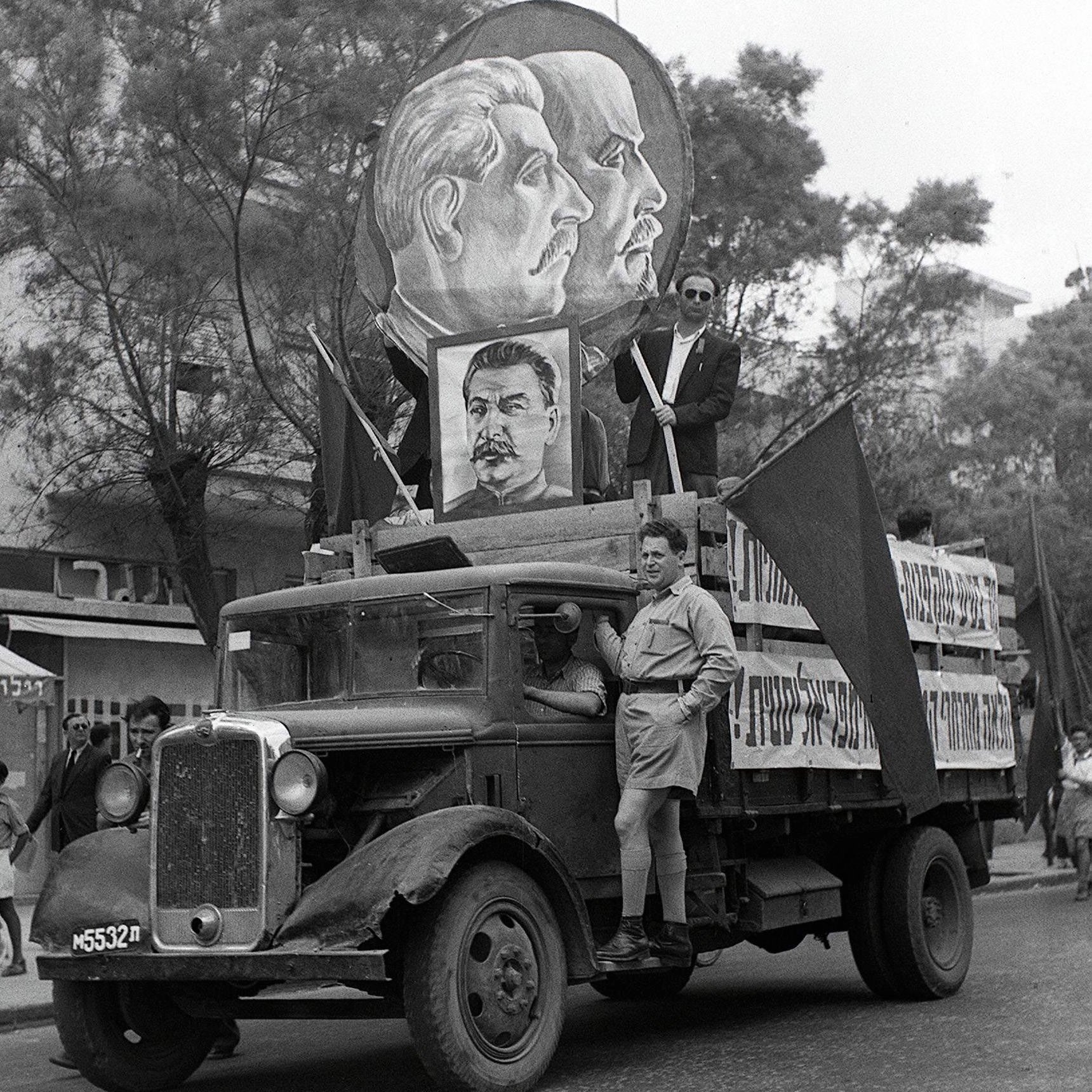 تصویر نشان‌دهنده یک خودروی قدیمی است که در یک مراسم راهپیمایی یا تظاهرات شرکت کرده است. بر روی ماشین بزرگ‌نمایی از چهره‌های افرادی با موهای کوتاه و مقدار زیادی نماد و پرچم‌هایی وجود دارد. فردی در حالت ایستاده بر روی خودرو قرار دارد و به نظر می‌رسد که در حال صحبت یا اشاره به تماشاگران است. پس‌زمینه شامل گروهی از مردم است که در کنار خیابان ایستاده‌اند، و فضایی از شور و اشتیاق را در این رویداد به تصویر می‌کشد.