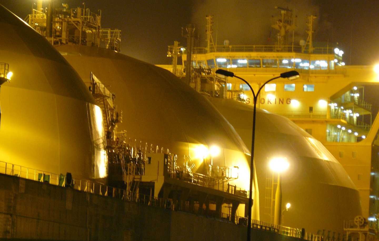 L'image montre une scène nocturne avec de grands réservoirs de stockage, probablement liés à l'industrie du gaz naturel. Les réservoirs ont des formes arrondies et sont éclairés par des lumières jaunes, créant une ambiance industrielle. En arrière-plan, on peut voir un navire, équipé de dispositifs de chargement ou de déchargement, suggérant une activité portuaire. La combinaison de l'illuminations et des structures métalliques donne une impression de travail intense et de technologie avancée.
