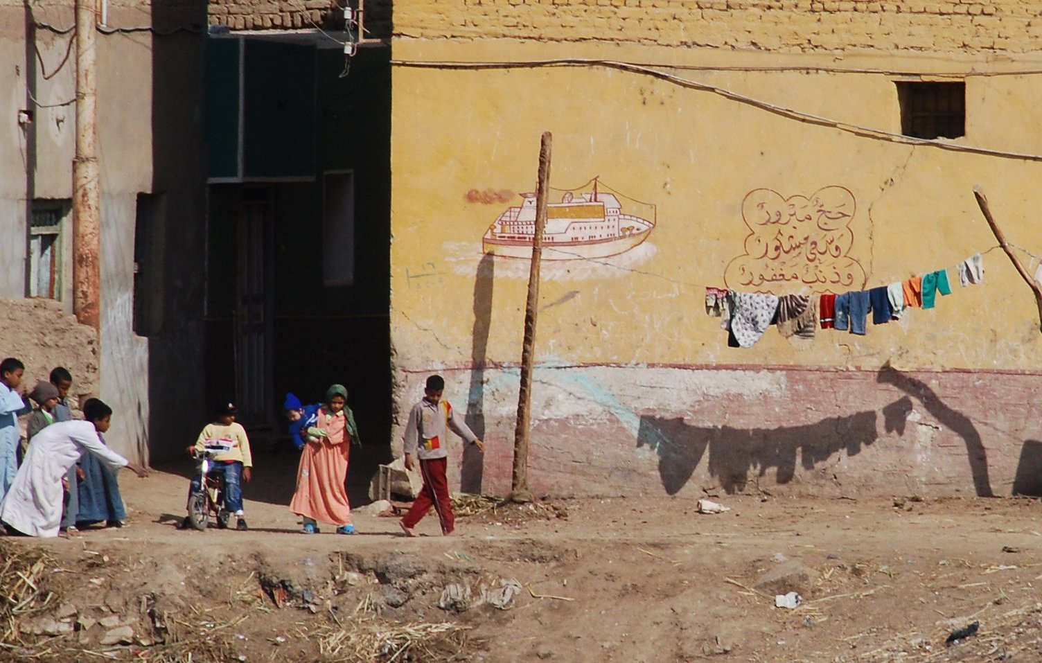 The image depicts a scene from a rural or semi-urban area, featuring a group of people interacting outside. A child in a wheelchair is being pushed forward by an adult, while two other children walk beside them. In the background, there is a building with a yellow wall that has some painted decorations or text. Above this, a clothesline displays various articles of clothing hanging to dry. The ground appears uneven, with some debris scattered around. Overall, the scene conveys everyday life in a community setting.