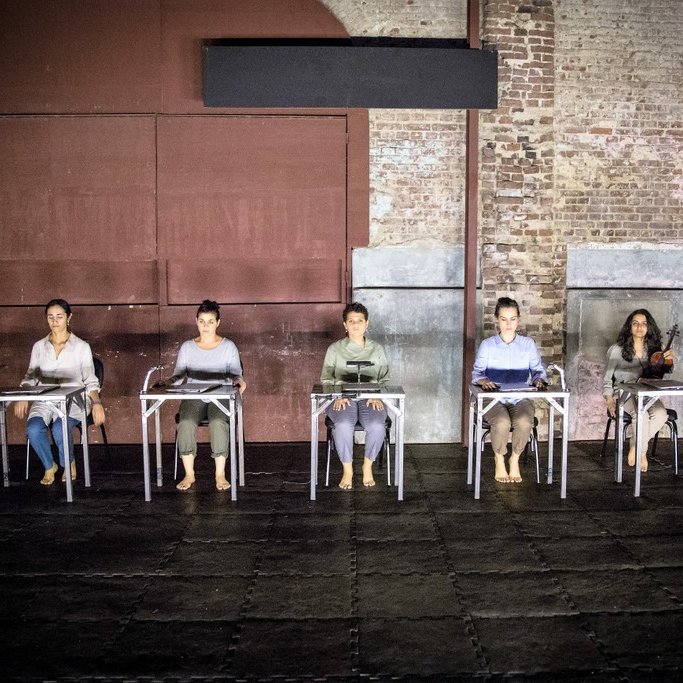 L'image montre un groupe de cinq personnes assises sur des chaises, disposées en ligne. Elles portent des vêtements simples et sont pieds nus, ce qui donne une impression de simplicité et d'intimité. L'environnement est industriel, avec des murs en brique apparente et une lumière tamisée qui crée une ambiance à la fois contemplative et moderne. Les personnes semblent concentrées, chacune avec un objet devant elle, suggérant un moment de performance ou de réflexion partagée.