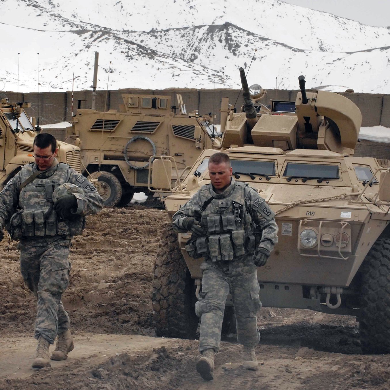 En la imagen se pueden ver a dos soldados caminando por un terreno irregular y arenoso. Están vestidos con uniformes militares y parecen estar en un entorno montañoso, con algunas unidades de vehículos blindados al fondo. La atmósfera es de actividad militar, con un paisaje que sugiere que están en una misión en un área remota. La nieve se puede observar en las montañas que rodean la escena.