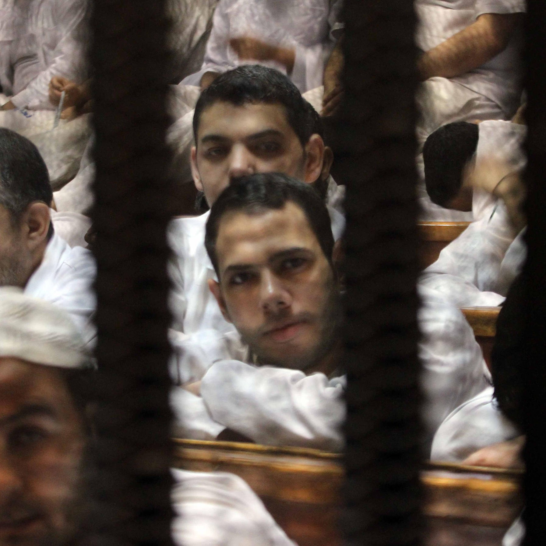 تظهر الصورة مجموعة من الرجال يجلسون في قاعة المحكمة، مع وجود قضبان أو شبك يظهر في المقدمة. يبدو أنهم يرتدون ملابس بيضاء ويظهر على وجوههم تعابير مختلفة. بعضهم ينظر إلى الكاميرا بينما يبدو آخرون مشغولين بأفكارهم. الجو العام يوحي بالتوتر والجدية، مما يعكس طبيعة المكان الذي يصور.