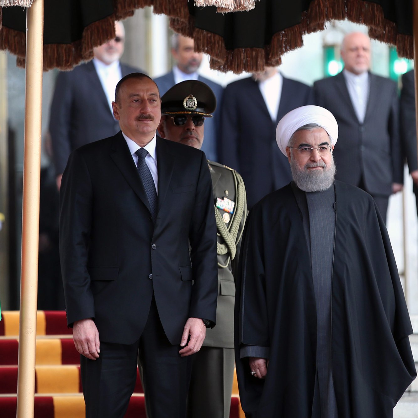En la imagen se pueden ver a dos líderes de países que están de pie juntos. A la izquierda, hay una bandera de Azerbaiyán. Uno de los líderes lleva un traje negro y corbata, mientras que el otro viste una indumentaria tradicional oscura. Detrás de ellos, hay un grupo de personas que parecen ser miembros de la seguridad o funcionarios, y en el fondo se observan algunos edificios. La escena parece ser un acto formal o una reunión oficial.