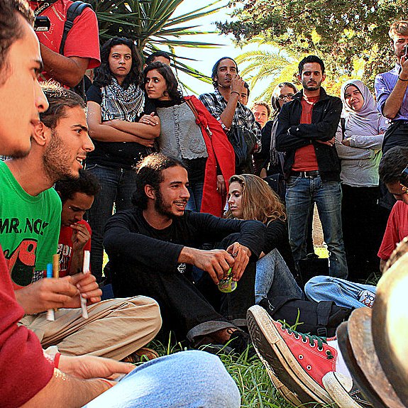 L'image montre un groupe de personnes rassemblées dans un espace extérieur, probablement un parc. Certains d'entre eux sont assis sur le sol, tandis que d'autres se tiennent debout ou sont légèrement en retrait. La scène semble conviviale et décontractée, avec des gens qui discutent, rient ou écoutent de la musique. Au centre, on aperçoit des instruments de musique, suggérant qu'il y a une performance musicale en cours. Les personnes présentes semblent diverses, avec des styles vestimentaires variés, ce qui indique une atmosphère informelle et communautaire.