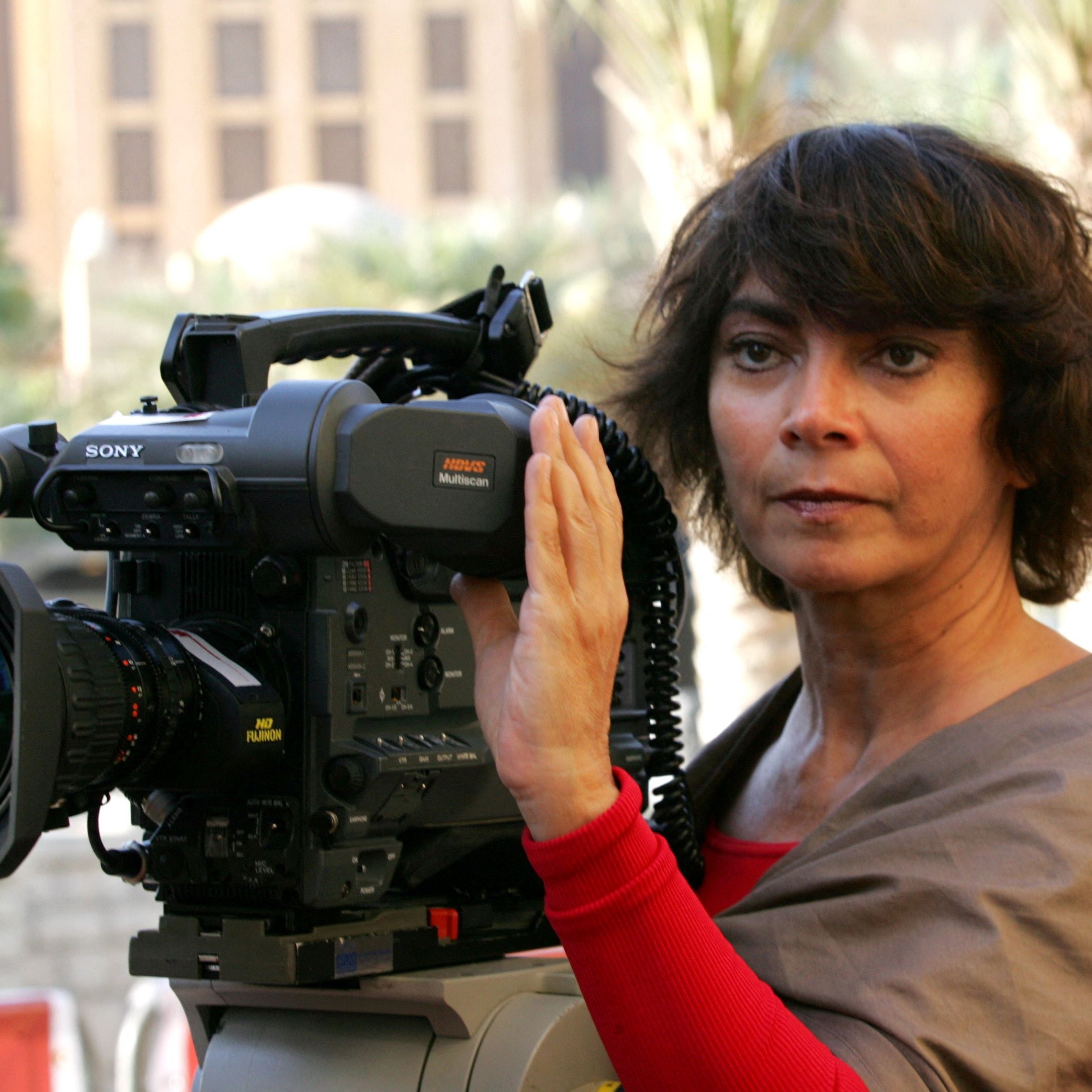 La imagen muestra a una mujer de cabello oscuro y rizado, que parece estar sosteniendo y operando una cámara de video profesional. Ella está mirando a la cámara con una expresión seria y concentrada. Viste una blusa color claro y tiene una capa o chal de un color más oscuro. Al fondo, se pueden ver edificios y un ambiente urbano. La escena sugiere que está en un evento o en el lugar de una grabación.