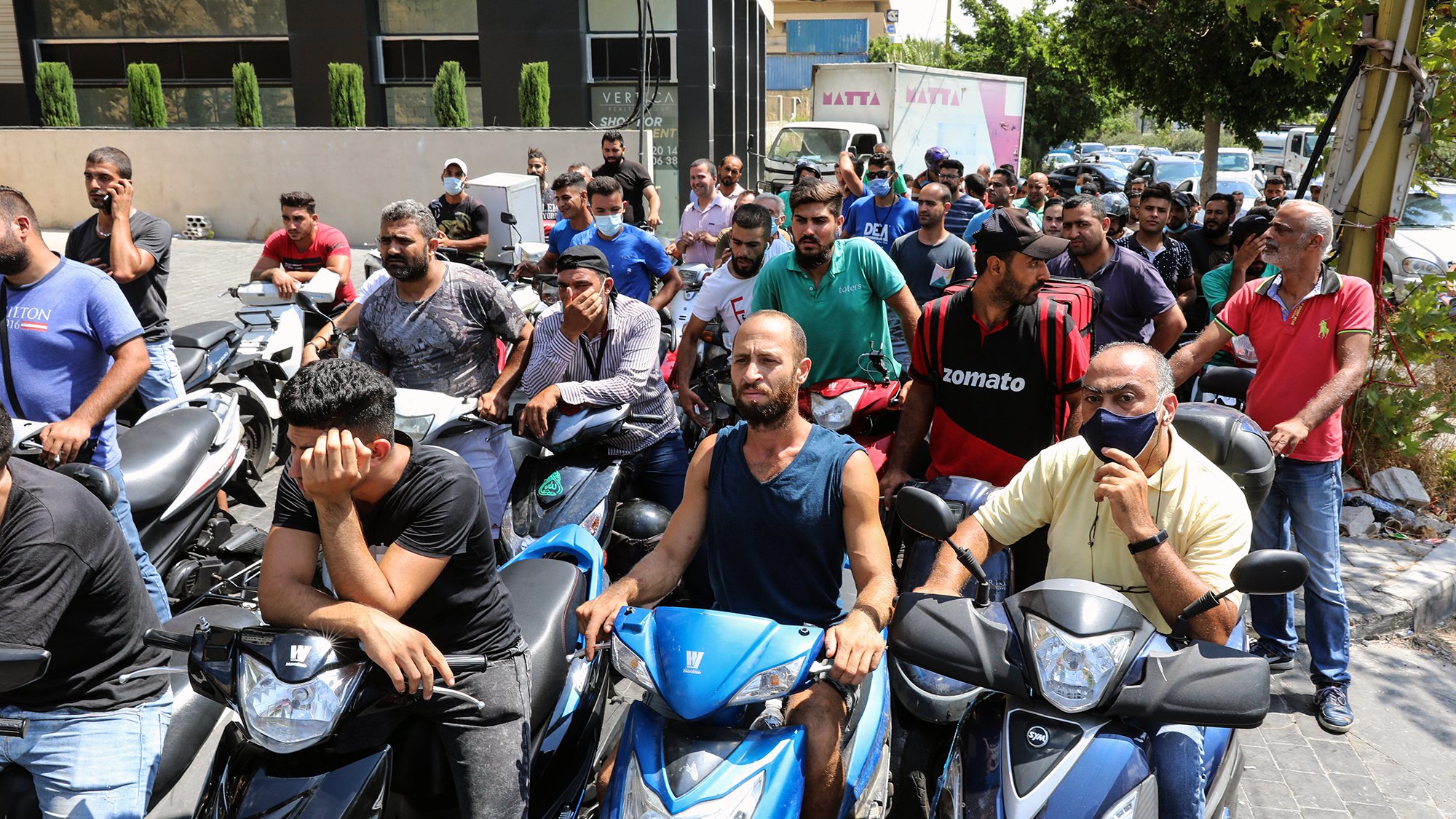 L'image montre un groupe de personnes, principalement des hommes, rassemblés sur des motos dans une file d'attente. Ils semblent attendre pour quelque chose, peut-être un service ou un approvisionnement. Certains affichent des expressions de frustration ou d'impatience. En arrière-plan, on peut voir des bâtiments et des arbres qui ajoutent au cadre urbain. L'atmosphère donne une impression de tension liée à une situation d'attente prolongée.