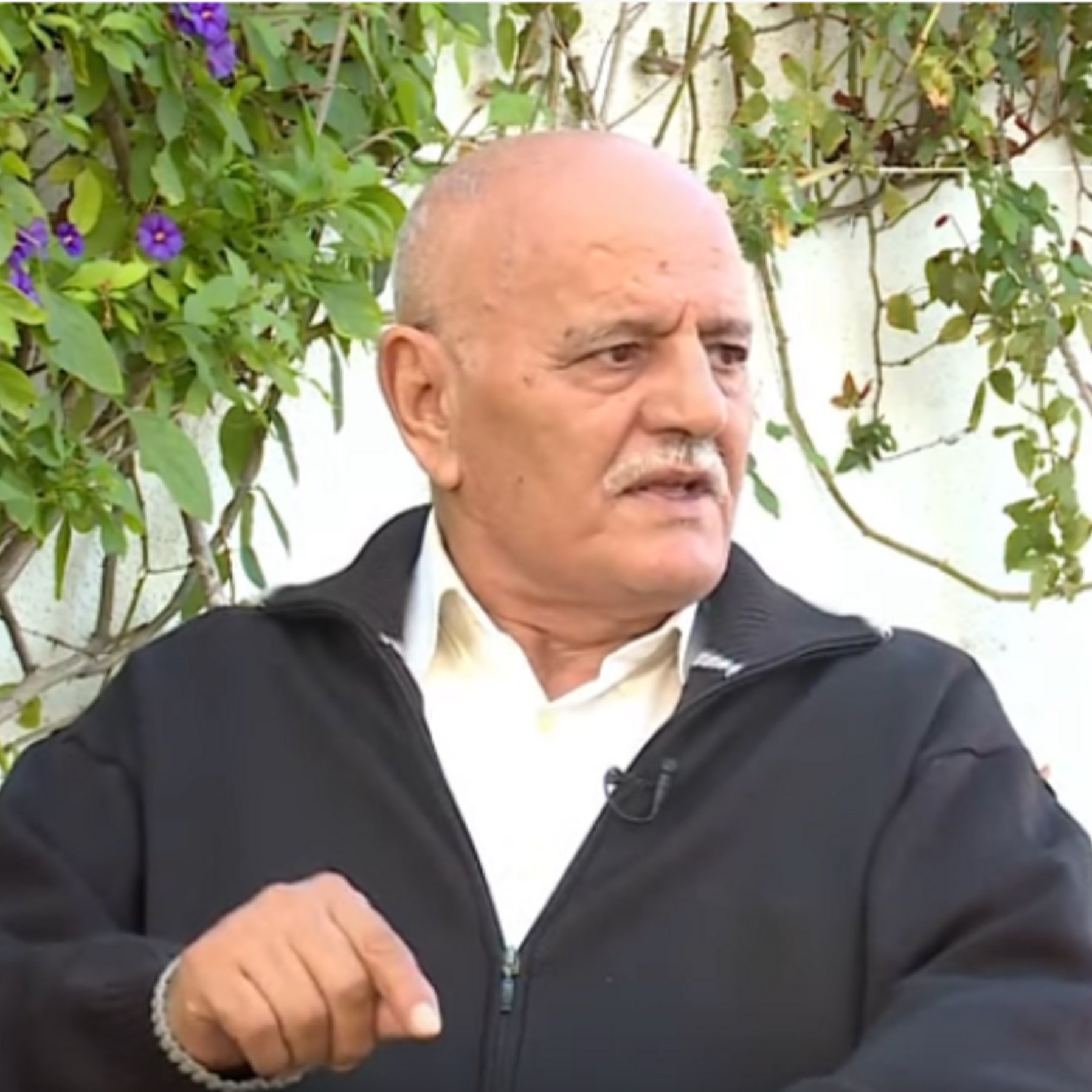 L'image montre un homme âgé assis en extérieur. Il porte une veste noire et semble être en train de s'exprimer. En arrière-plan, on peut voir des plantes et des fleurs, ce qui donne une ambiance naturelle à la scène. Son expression faciale et son geste avec la main suggèrent qu'il est en train de parler d'un sujet important ou d'expliquer quelque chose.