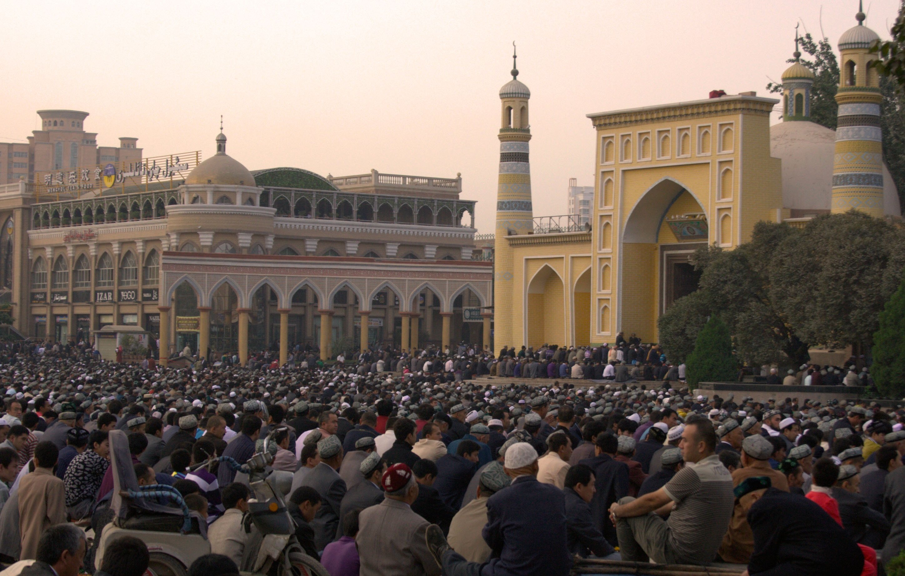 تش depicts الصورة تجمعًا كبيرًا من الناس، حيث يجلس عدد كبير من الأفراد في ساحة مفتوحة. في الخلفية، يمكن رؤية معمار تقليدي يتضمن مآذن وقباب، مما يشير إلى أنه قد يكون موقعًا دينيًا. السماء في حالتها النهارية تكتسي بألوان دافئة، مما يضفي جواً خاصاً على المشهد. يبدو أن الناس مجتمعون لأداء نشاط جماعي، ربما صلاة أو احتفال.