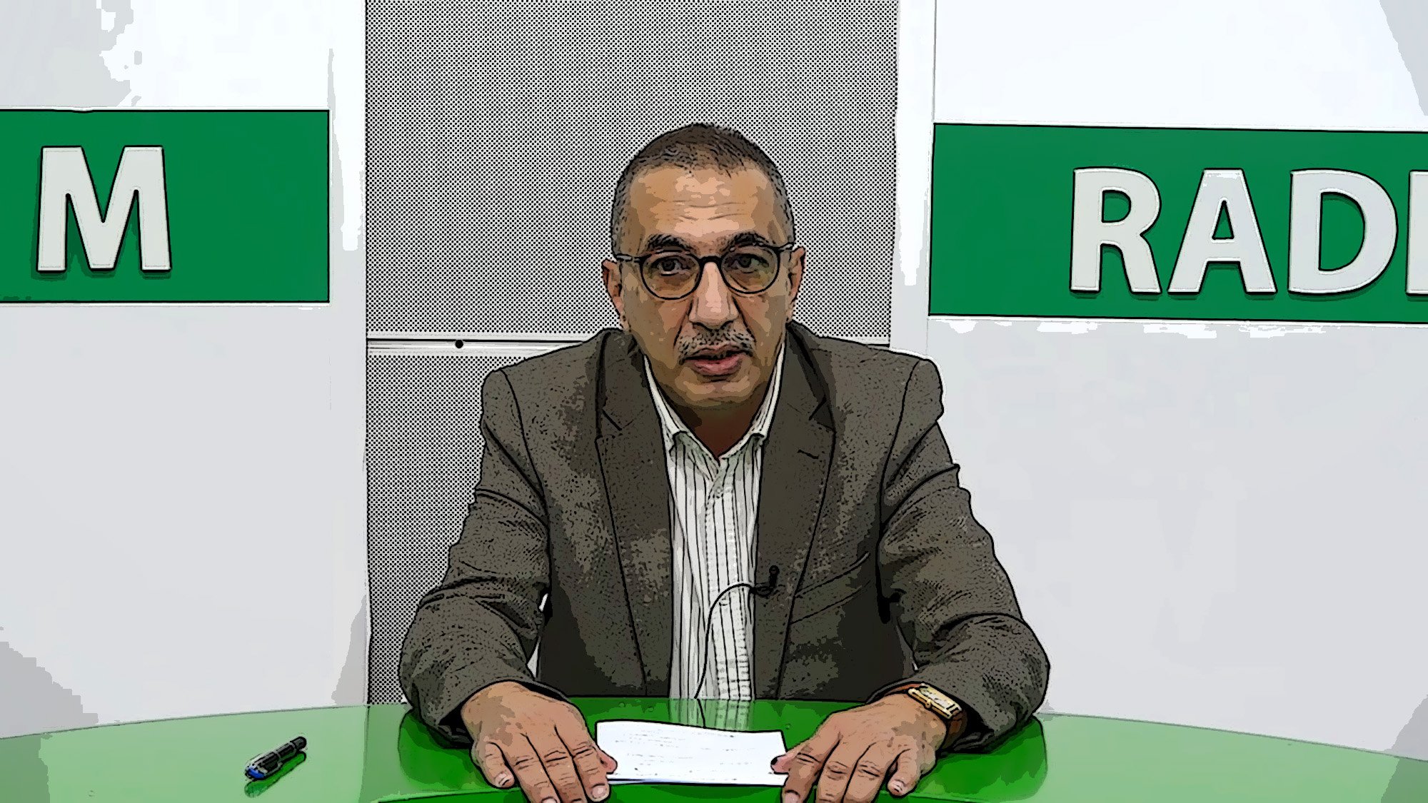 L'image montre un homme devant un bureau, probablement dans un studio de télévision ou de radio. Il porte des lunettes et un costume, avec une chemise rayée. Le bureau est vert, et il semble tenir des documents ou un script. En arrière-plan, il y a des éléments graphiques avec les mots "RADIO" et une autre inscription non visible. L'homme a une expression sérieuse et semble prêt à faire une présentation ou une annonce.