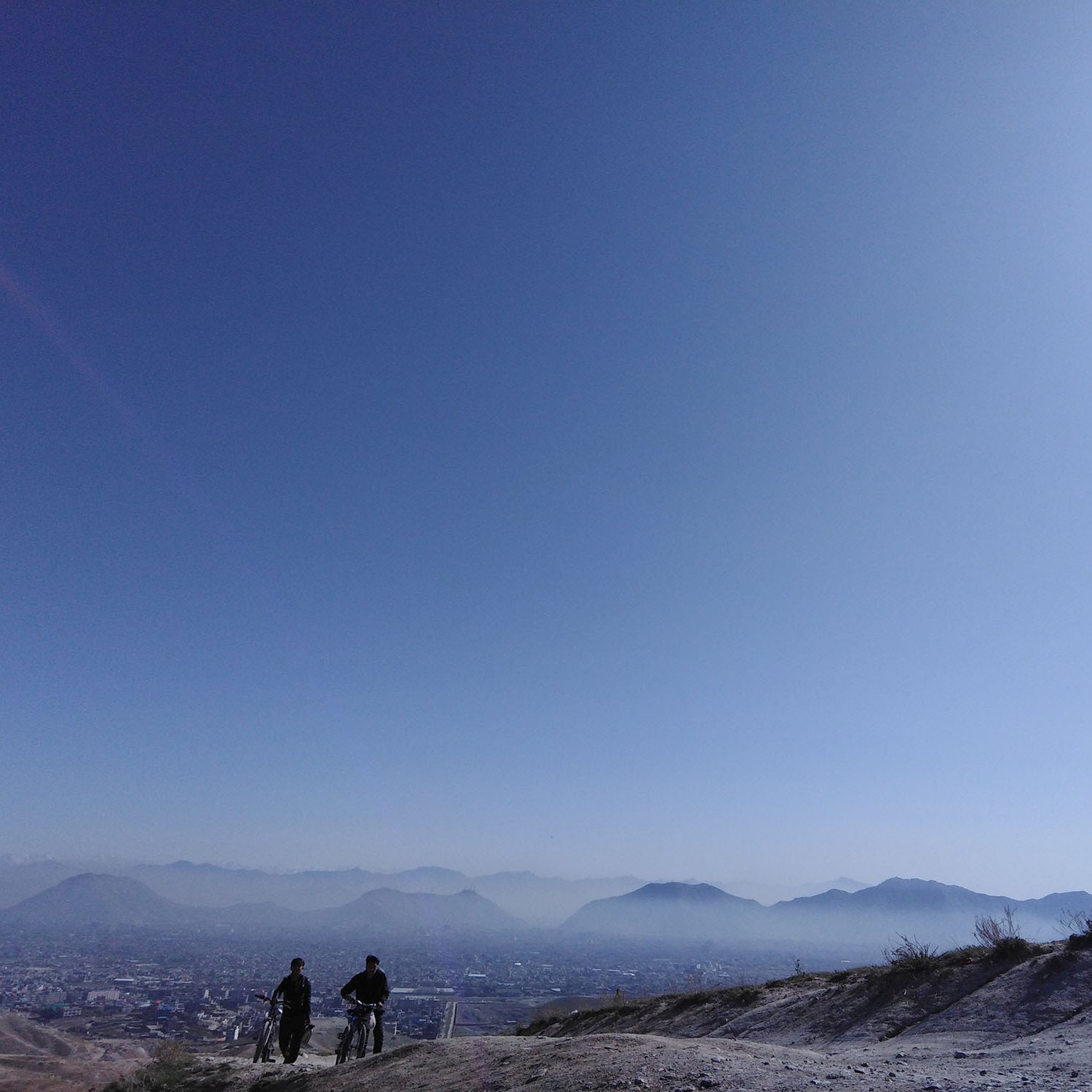 The image depicts a serene landscape with two individuals walking along a trail on a hillside. They are silhouetted against a vast, clear blue sky, which suggests a sunny day. In the background, there are distant mountains and a sprawling cityscape, partially visible below. The scene captures a sense of tranquility and openness, highlighting the natural beauty of the setting.