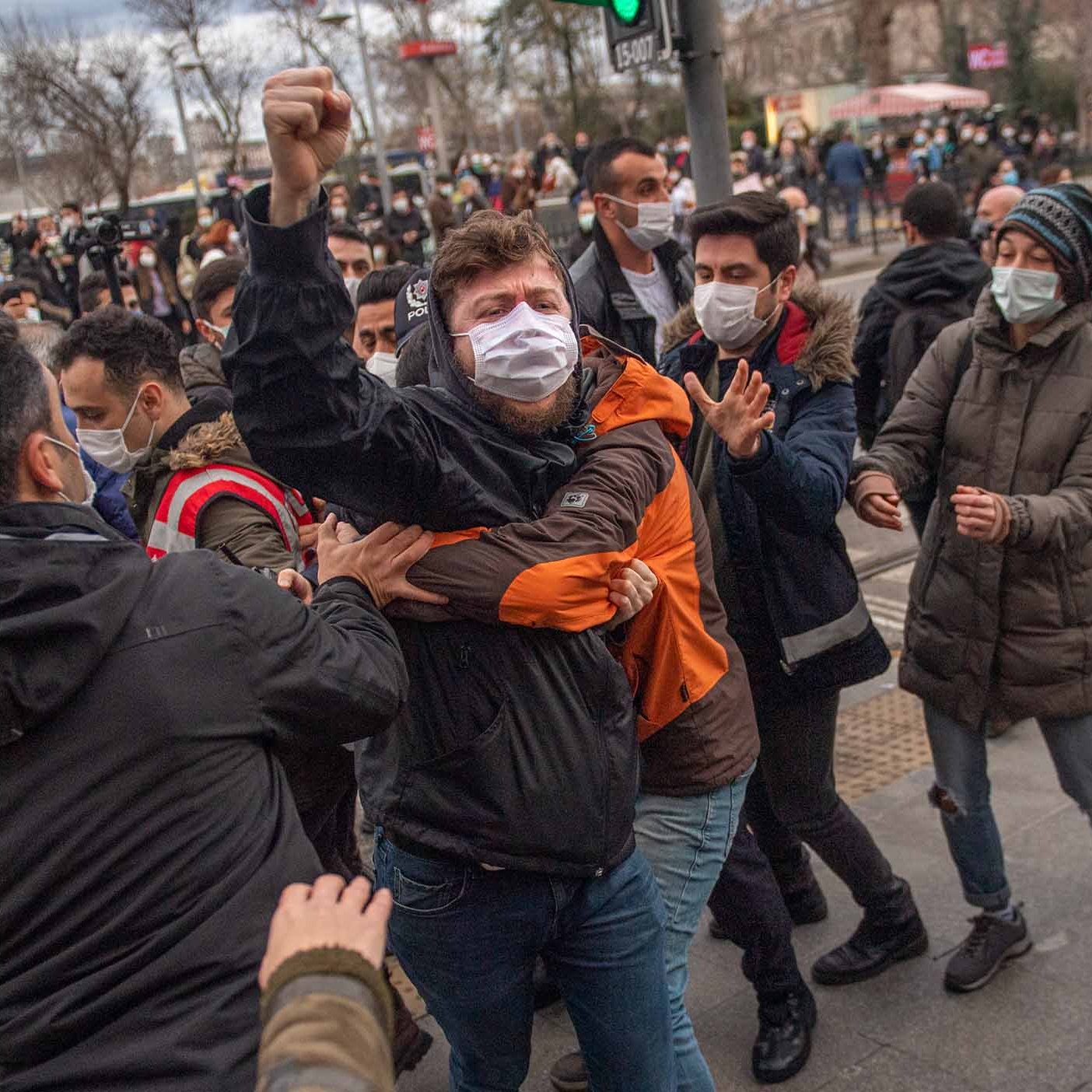 تظهر الصورة مجموعة من الأشخاص في مشهد حيوي، حيث يبدو أن هناك احتجاجًا أو تجمعًا. بعضهم يرتدي الكمامات، مما يشير إلى الالتزام بإجراءات السلامة. في المقدمة، يُلاحظ شاب يرفع يده في إشارة تعبيرية، بينما يتداخل الآخرون حوله، مما يعكس حالة من التوتر أو الحماس. الخلفية تضم تفاصيل حضرية، مثل إشارات المرور والمباني، مما يوحي بأن المكان قد يكون في مدينة كبيرة.