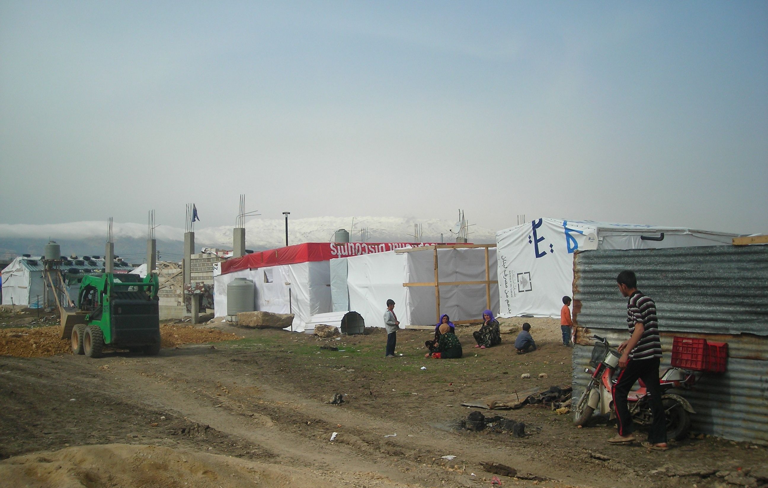 L'image montre un camp de réfugiés ou de personnes déplacées, avec des tentes blanches et des structures temporaires. Il y a un tracteur ou un véhicule de service vert à gauche. Plusieurs personnes sont visibles dans le camp, certaines assises ou debout près des tentes. Le terrain est poussiéreux, et en arrière-plan, on peut apercevoir des montagnes ou des collines. L'ensemble du paysage semble austère et reflète les conditions de vie des occupants. L'atmosphère paraît tranquille, bien que l'environnement soit rudimentaire.