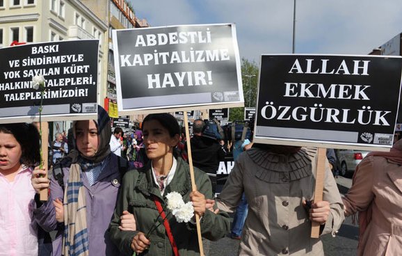 L'image montre une manifestation avec des personnes portant des pancartes. Les manifestants semblent exprimer des revendications contre la pression et l'oppression, en mettant en avant des messages en turc. Certaines femmes portent des foulards, suggérant un contexte culturel spécifique. On peut sentir une atmosphère de mobilisation collective.