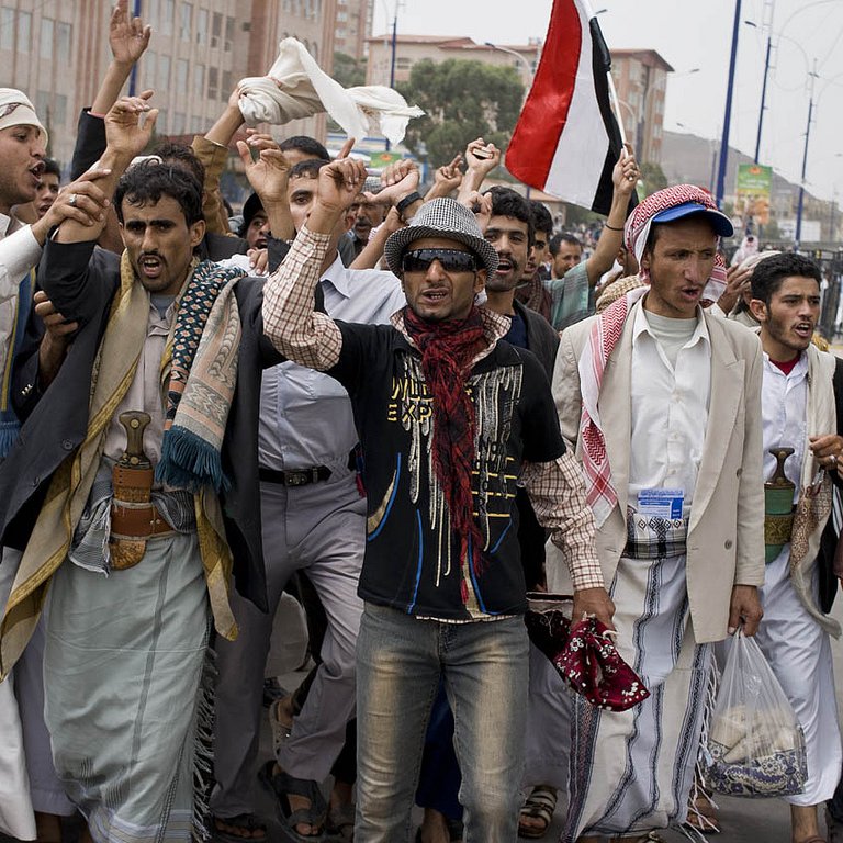 L'image montre un groupe de personnes en train de manifester dans une rue. Elles semblent exprimer des revendications, levant les bras et brandissant des drapeaux, notamment un drapeau rouge. Les manifestants portent des vêtements traditionnels yéménites, avec divers styles de vêtements. L'ambiance paraît énergique et engagée, comme une expression de protestation ou de soutien à une cause.