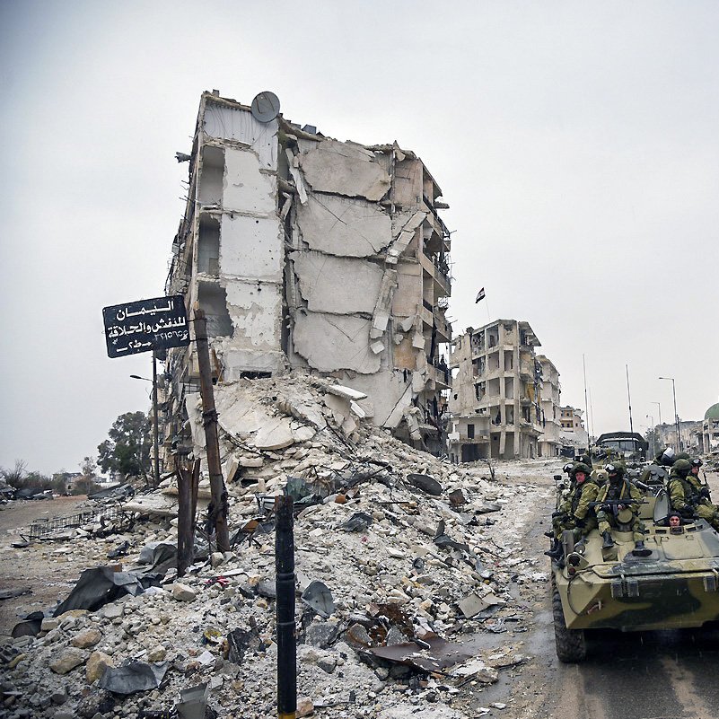 تصویر نشان‌دهندهٔ یک منطقهٔ ویران شده است که در آن چند ساختمان تخریب شده و در حال فرو ریختن به نظر می‌رسند. در این صحنه، یک وسیله نقلیه نظامی در حال حرکت است و تعدادی سرباز در آن نشسته‌اند. بقیهٔ محیط شامل زباله‌ها و نشانه‌های ویرانی است که به وضعیت بحرانی منطقه اشاره دارد. هوا ابری و تیره به نظر می‌رسد و فضایی خالی و ساکت را ایجاد کرده است.