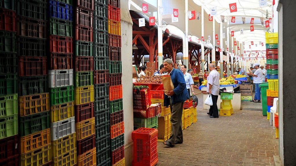 L'image montre un marché couvert animé, avec des étals bien organisés. On peut y voir des rangées de caisses colorées, de différentes tailles et en plastique, empilées de manière ordonnée. Des personnes se déplacent dans le marché, certaines portant des sacs, tandis que d'autres semblent inspecter les produits disponibles. Le tout est sous une structure avec un toit, permettant de se préserver du soleil. Des drapeaux tunisiens décorent le plafond, ajoutant une touche locale à l'ambiance du lieu.