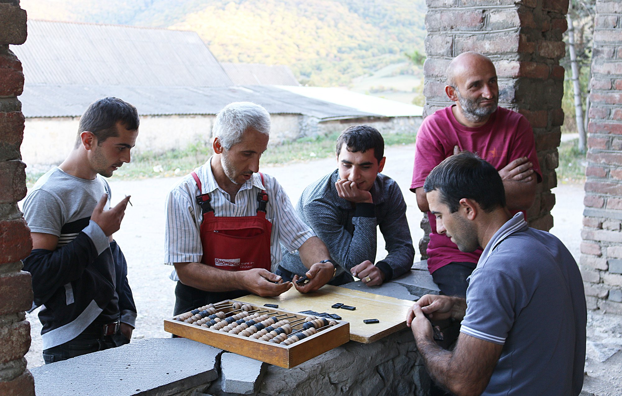 L'image montre un groupe de cinq hommes rassemblés autour d'une table en pierre en extérieur. Sur la table, il y a un plateau avec des pièces de jeu, et un homme au centre semble expliquer ou démontrer quelque chose. Les autres hommes écoutent attentivement, certains les bras croisés et d'autres penchés vers la table. Le décor naturel en arrière-plan présente des collines verdoyantes, ce qui suggère un environnement rural. L'atmosphère semble conviviale et détendue.