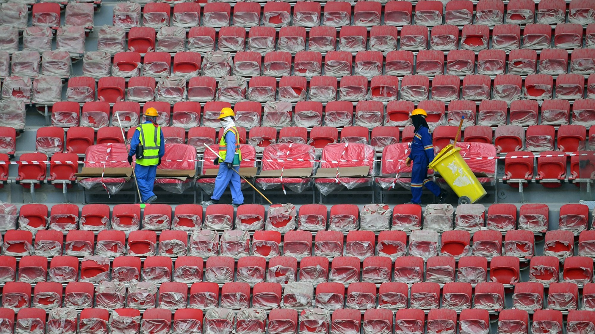 L'image montre des travailleurs dans un stade vide. Ils portent des casques de protection et des gilets réfléchissants. Les sièges du stade sont recouverts de plastique, et les travailleurs semblent être en train de nettoyer ou de préparer les lieux. On peut voir des rangées de sièges rouges. L'atmosphère paraît calme et organisée.