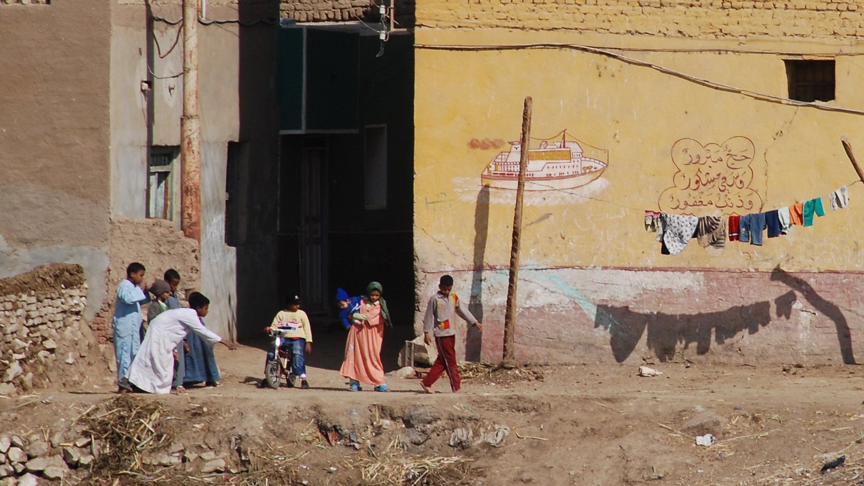 تظهر في الصورة مشهد لحياة يومية في منطقة شبه ريفية. في الجهة اليسرى يوجد شخص يرتدي ملابس بيضاء يحمل حقيبة، ويبدو أنه يسير نحو مجموعة من الأطفال والنساء. الأطفال يبدو أنهم يلعبون أو يتجهون إلى مكان ما. في الخلفية، توجد جدران ملونة تحتوي على رسوم جرافيتية وبعض الملابس المعلقة على حبل. الأرض تبدو جافة، مع بعض الحجارة والنباتات الصغيرة. يظهر كل ذلك الحياة البسيطة والمجتمع النشط في هذه المنطقة.