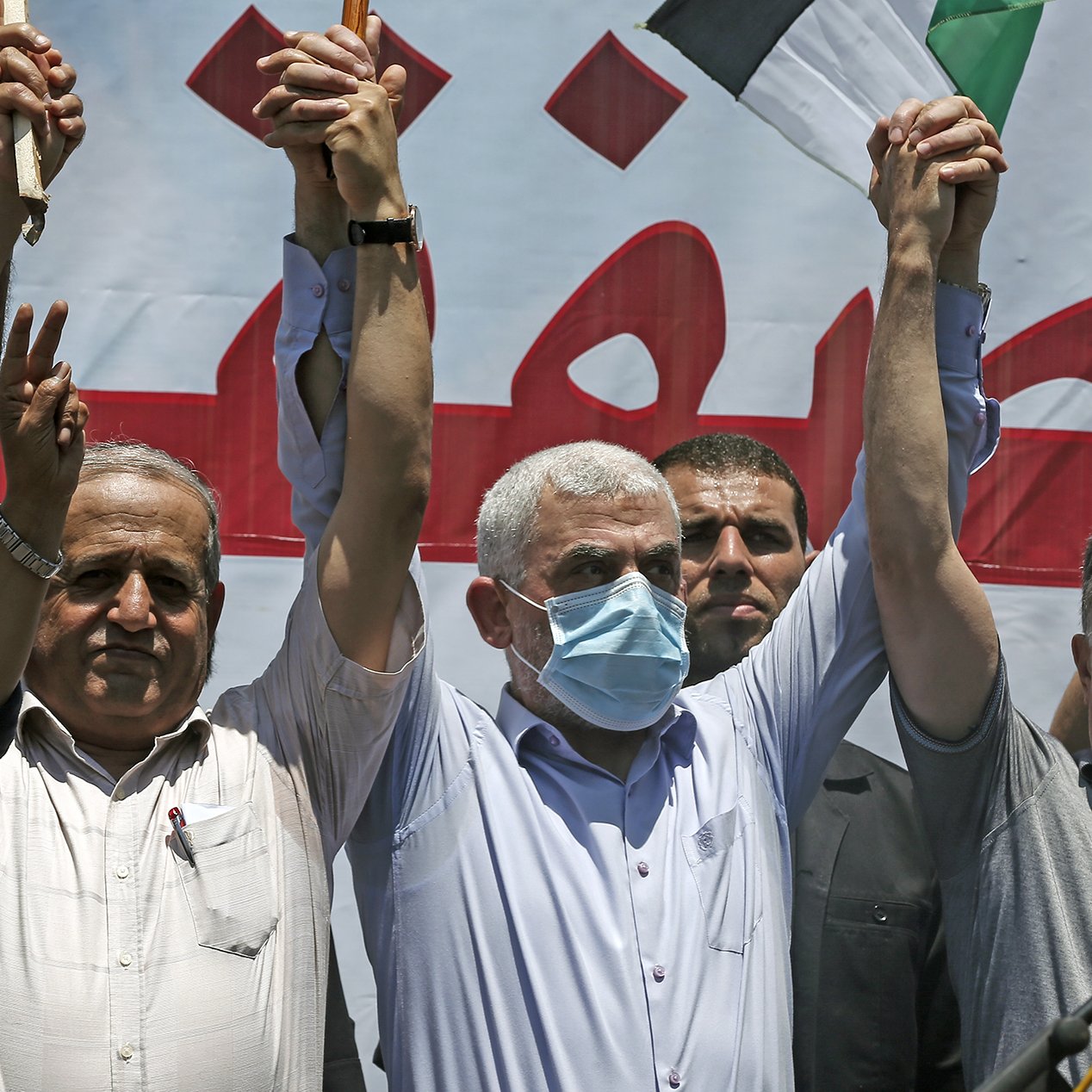 En la imagen se puede ver a un grupo de hombres en una manifestación. Ellos levantan las manos, mostrando un gesto de unidad o celebración. Uno de los hombres en el centro lleva una máscara facial, lo que sugiere que la situación se desarrolla en un contexto donde se requieren medidas de salud. Detrás de ellos hay una pancarta o cartel grande, con texto en árabe y un fondo que parece estar relacionado con la causa por la que están protestando. La atmósfera parece ser de fervor y apoyo colectivo.