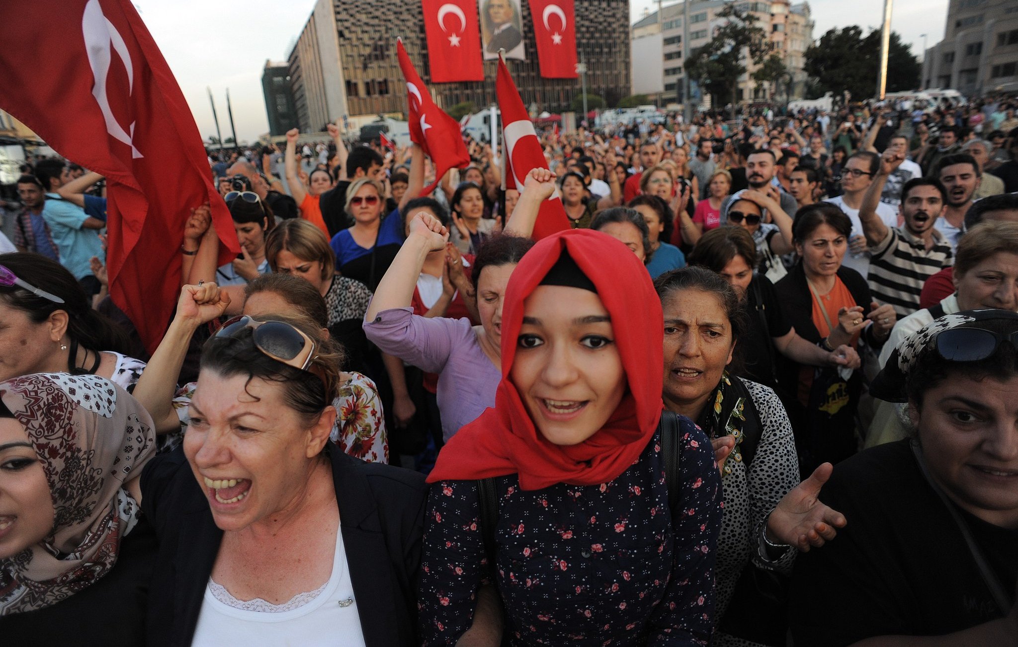 The image depicts a large crowd gathered, likely in an outdoor setting, with many individuals holding Turkish flags. People appear to be passionately engaged, with expressions of enthusiasm and solidarity. Some individuals are wearing traditional attire, while others wear modern clothing. The background features a building adorned with large banners, adding to the sense of national pride and collective emotion in the scene. The atmosphere seems charged with energy, suggesting a demonstration or celebration of national significance.