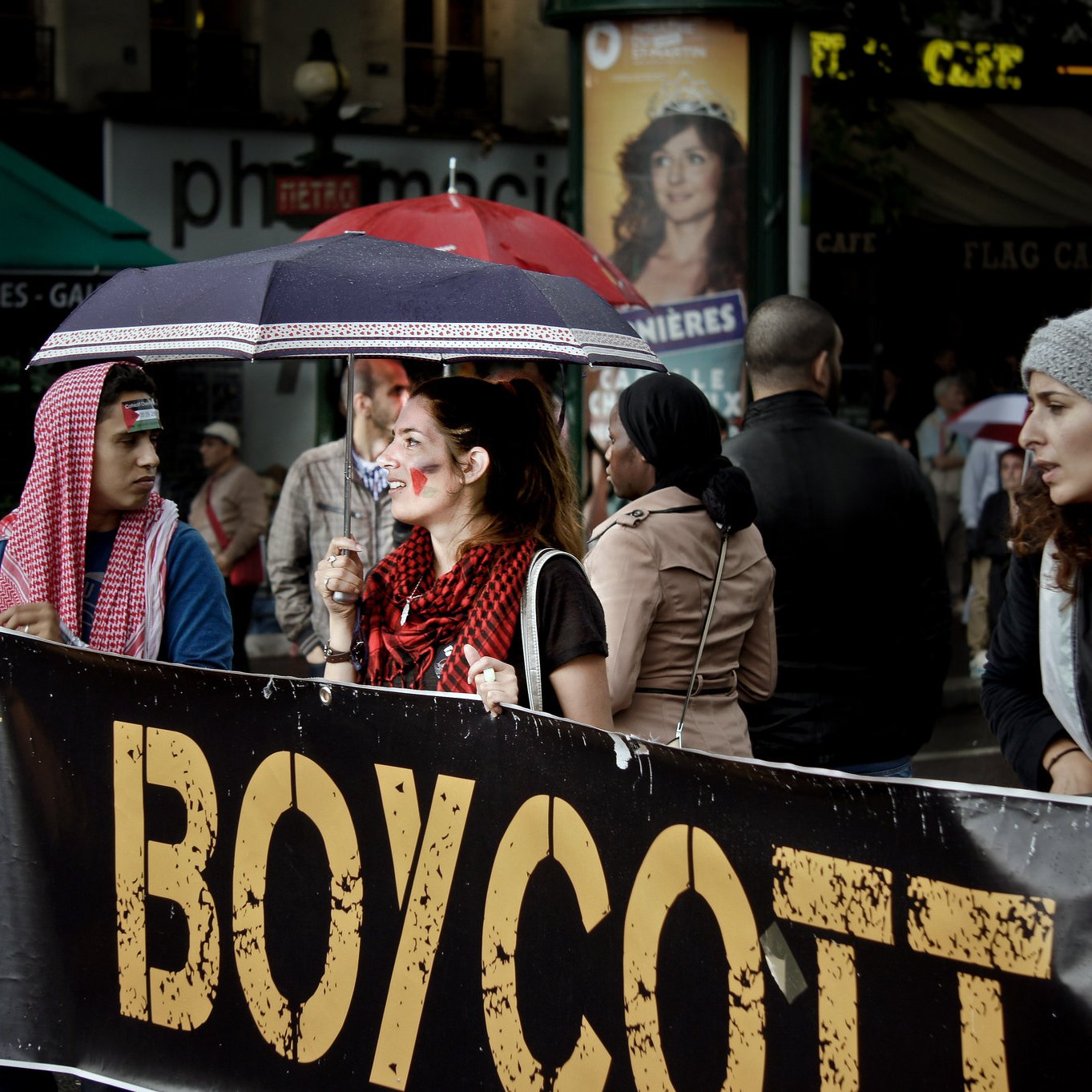 L'image montre un groupe de personnes participant à une manifestation. Au premier plan, on voit une femme tenant un grand panneau qui dit "BOYCOTT". Elle est entourée de plusieurs manifestants, dont certains portent des accessoires ou des vêtements aux couleurs de différents pays. Il y a aussi des parapluies, probablement à cause de la météo. L'ambiance semble engagée, avec un mélange de détermination et de solidarité parmi les participants.