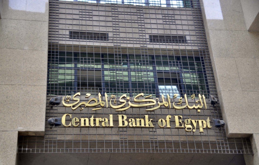 L'image montre la façade d'un bâtiment moderne, probablement celle de la Banque centrale d'Égypte. On peut voir le nom de l'institution écrit en arabe et en anglais, avec un design sobre et élégant. La structure est faite de granite et présente des grilles métalliques sur les fenêtres, ce qui donne une impression de sécurité et de solidité.