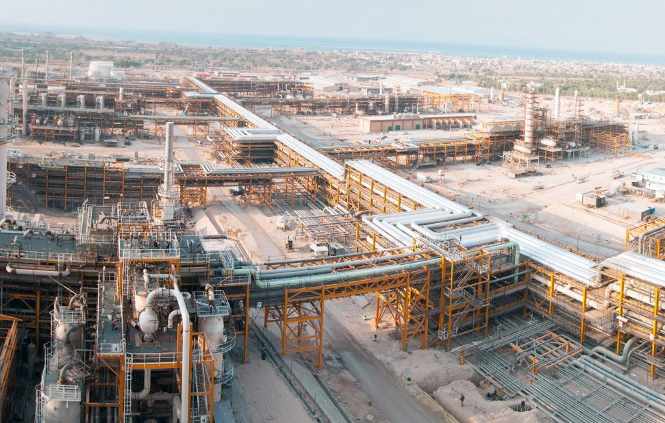 الصورة تُظهر مشهدًا واسعًا لمجمع صناعي، حيث تظهر مرافق ومعدات ضخمة. يمكن رؤية أنابيب كبيرة تمتد عبر الموقع، بالإضافة إلى أفران ومنشآت أخرى تشير إلى نشاط صناعي مكثف. الخلفية تتضمن المساحات المفتوحة والمباني التي تدل على طبيعة العمل في هذا المكان. السماء تبدو صافية، مما يضفي طابعًا مشمسًا على الصورة.