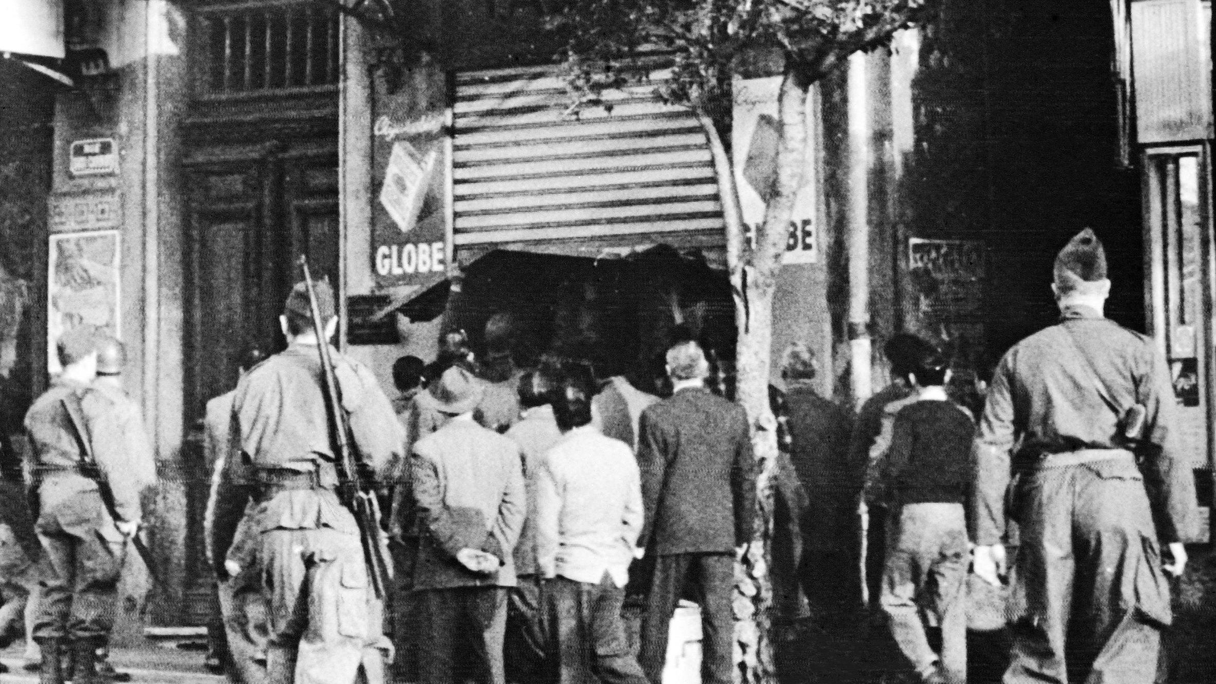 L'image montre une scène en noir et blanc où plusieurs soldats, en uniforme, se tiennent en arrière-plan. Devant eux, un groupe d'hommes en civil semble observer une situation à l'entrée d'un magasin ou d'un bâtiment, probablement une affaire en cours. La présence des soldats suggère un contexte de tension, peut-être lié à des événements historiques. Les détails de l'environnement, tels que les enseignes et l'architecture, évoquent une atmosphère urbaine, mais l'image elle-même semble figée dans le temps, ce qui renforce le caractère historique de la scène.