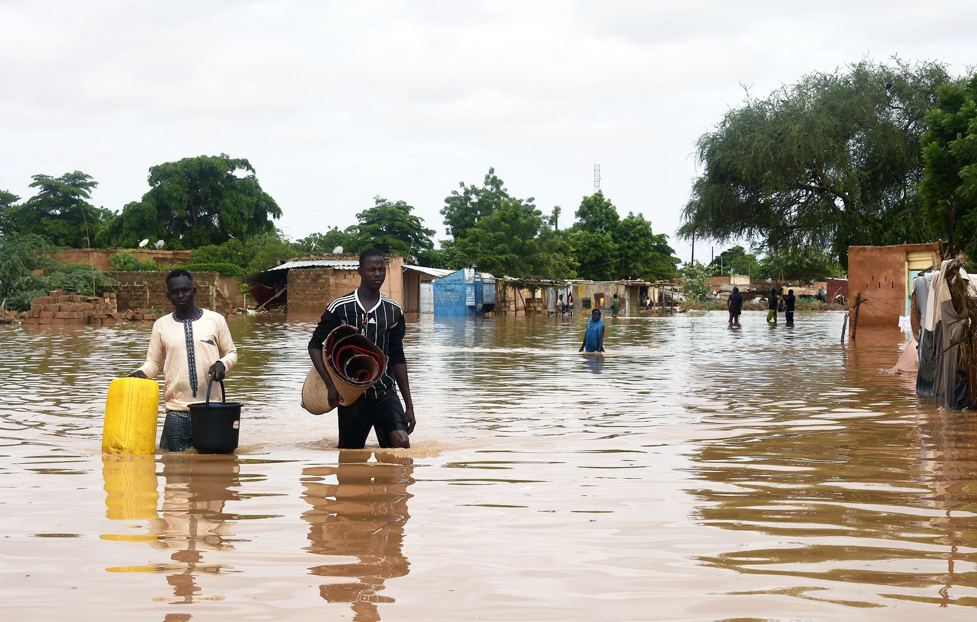The image depicts a flooded area, where individuals are walking through standing water. Two men are central in the foreground; one is carrying a bucket, while the other holds a rolled-up mat. In the background, makeshift shelters or homes are partially submerged, surrounded by trees and greenery. The scene reflects the impact of flooding, with water covering the ground and affecting the surrounding environment. The sky is overcast, suggesting ongoing adverse weather conditions.