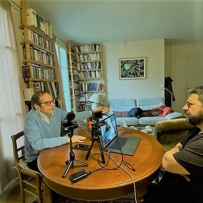The image depicts two men engaged in a conversation at a round wooden dining table. One man, wearing a light blue sweater and glasses, appears to be animatedly speaking and gesturing, while the other man, dressed in a black shirt, is listening attentively. On the table, there are microphones and a laptop, suggesting they might be recording a podcast or conducting an interview. In the background, a cozy living space is visible with shelves full of books and a sofa where another person seems to be resting. The overall atmosphere feels informal and collaborative.