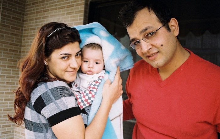 Sur cette image, on voit un couple et un bébé. La femme, à gauche, sourit et tient le bébé enveloppé dans une couverture, tandis qu'un homme, à droite, regarde la caméra en souriant également. Ils se tiennent tous les trois dans un environnement intérieur, probablement chez eux, avec un mur en briques en arrière-plan. L'atmosphère générale est chaleureuse et affectueuse.