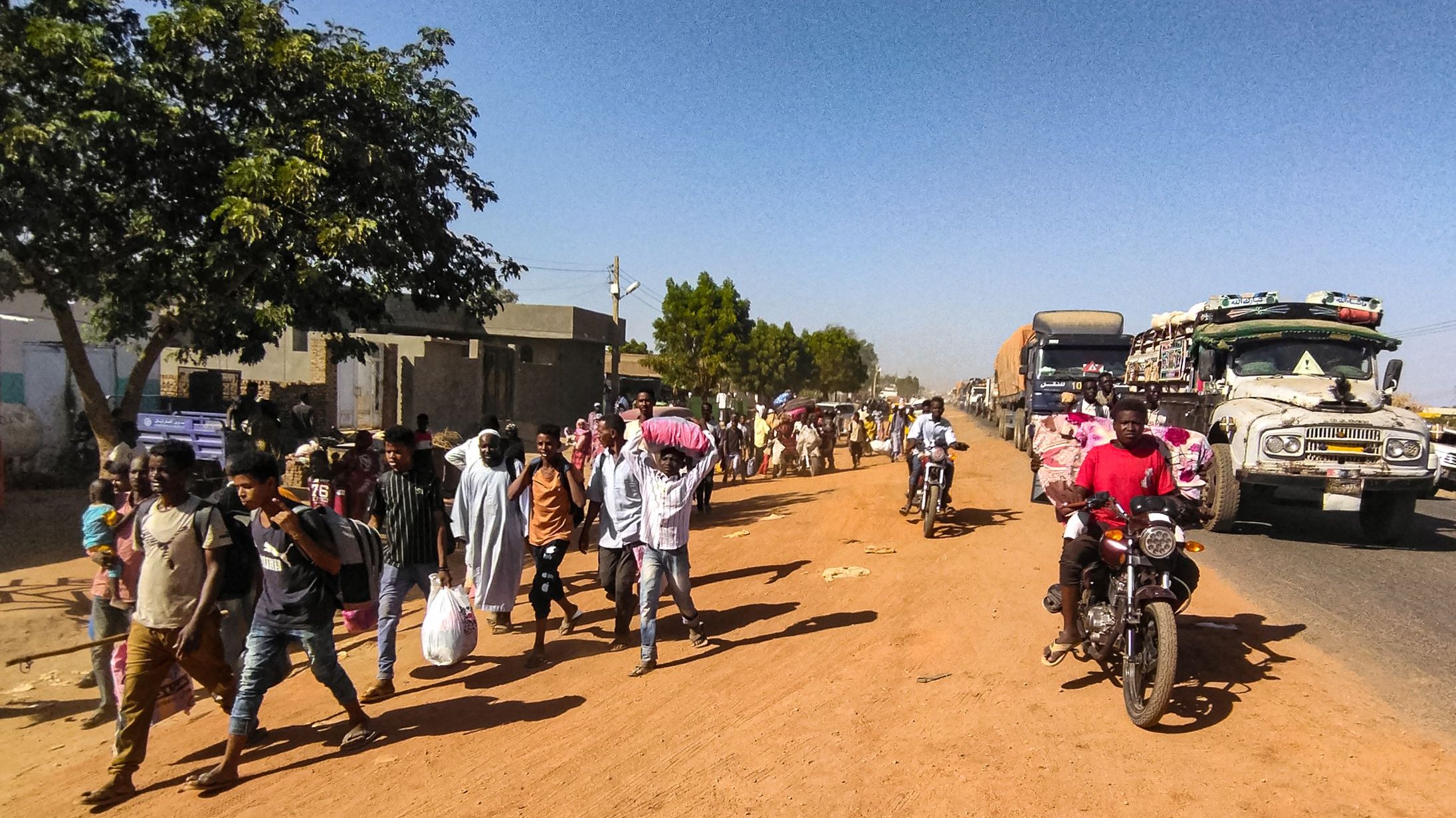 L'image montre une scène de rue animée dans une zone urbaine. Sur le côté gauche, on voit un arbre et des bâtiments, tandis que des gens marchent le long de la route en terre. Parmi eux, certains portent des sacs ou des charges sur la tête. Des véhicules, y compris des camions et des motos, circulent sur la route, créant une ambiance dynamique. L'éclairage semble être en plein jour, suggérant une activité quotidienne typique dans cette communauté.