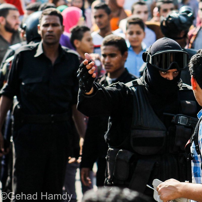 L'image montre une scène de confrontation dans un contexte urbain. À l'avant, un homme vêtu d'un uniforme noir, portant un casque et une visière, semble interagir avec un autre homme qui porte une chemise à carreaux. Ce dernier semble questionné ou arrêté, tandis qu'un groupe de personnes les entoure, observant la situation. On peut percevoir une atmosphère tendue, avec des expressions variées sur les visages des spectateurs. La scène suggère un moment de tension ou de dispute, probablement lié à une manifestation ou à une intervention policière.