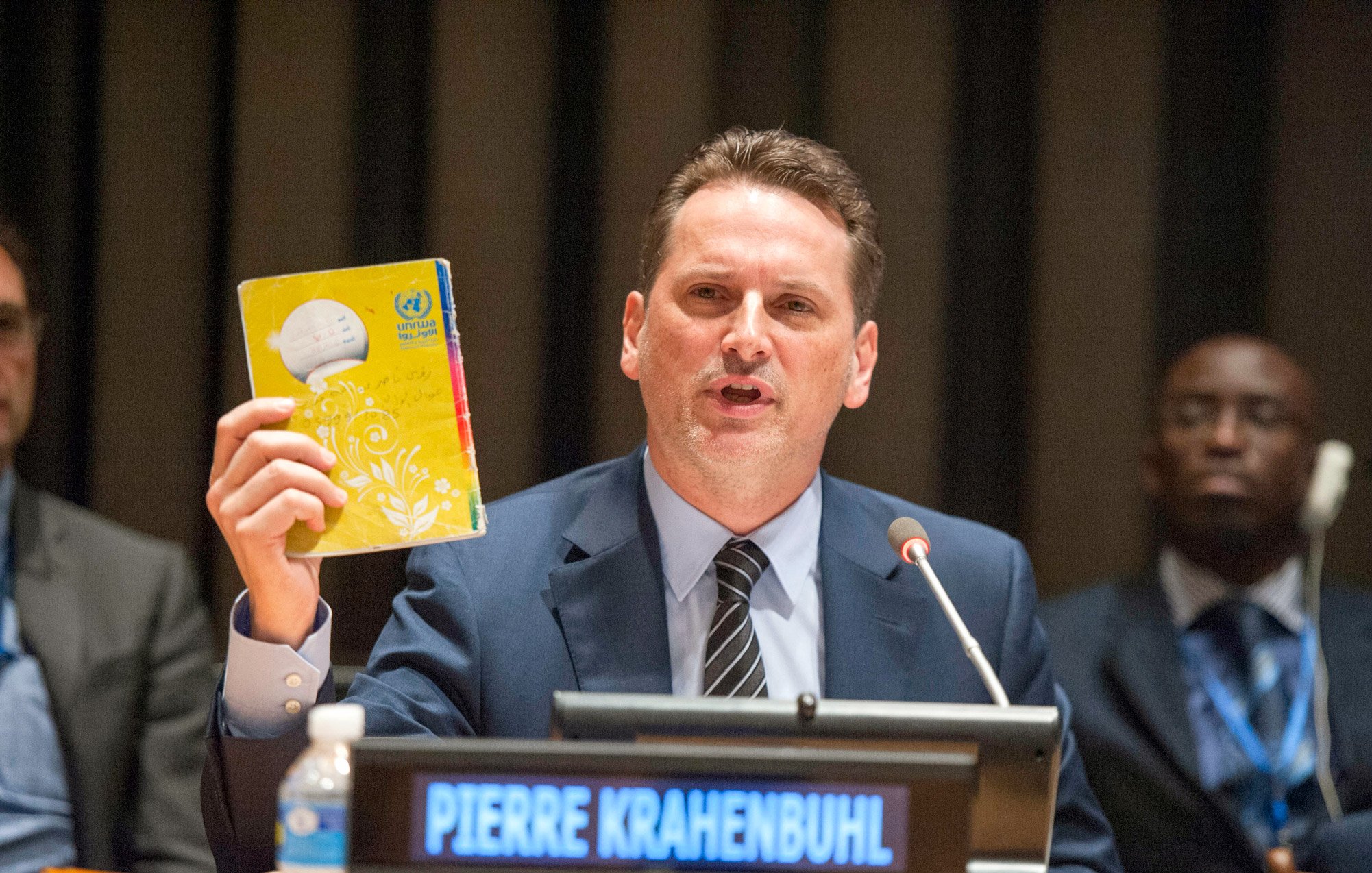 The image shows a man speaking at a podium during a conference or meeting, likely at a United Nations event. He is holding up a yellow booklet or document, which appears to be significant to his presentation. The nameplate in front of him reads "PIERRE KRAHENBUHL." In the background, there are several other individuals seated, and the setting has a formal atmosphere typical of international discussions. The man is engaged and emphasizing a point while addressing the audience.